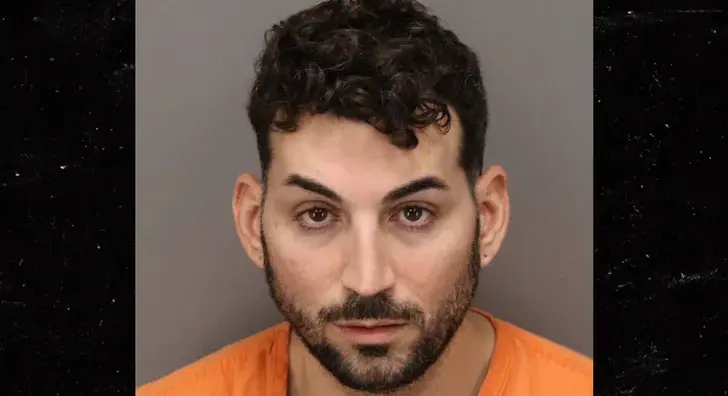 A man with short curly hair and a beard, wearing an orange shirt, in a mug shot.