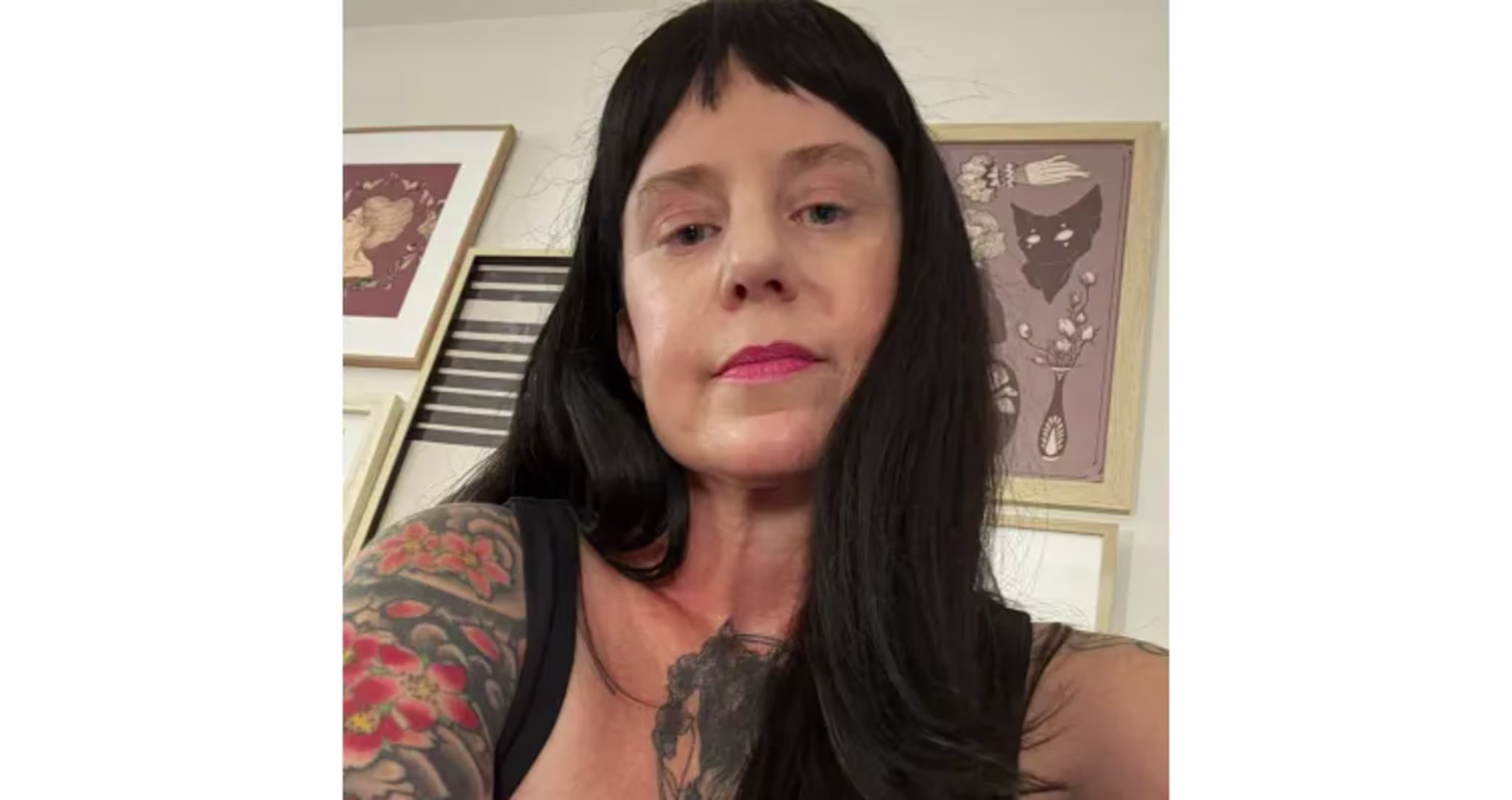 Rebecca Luna with long black hair and tattoos on their arm and chest, wearing a black top, in a room with framed art on the wall.