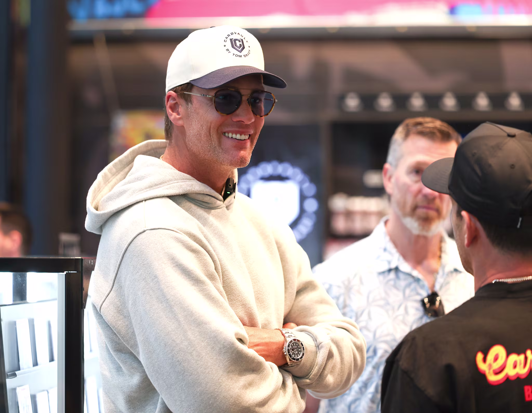 A man in a white cap and sunglasses smiles while talking to others indoors.