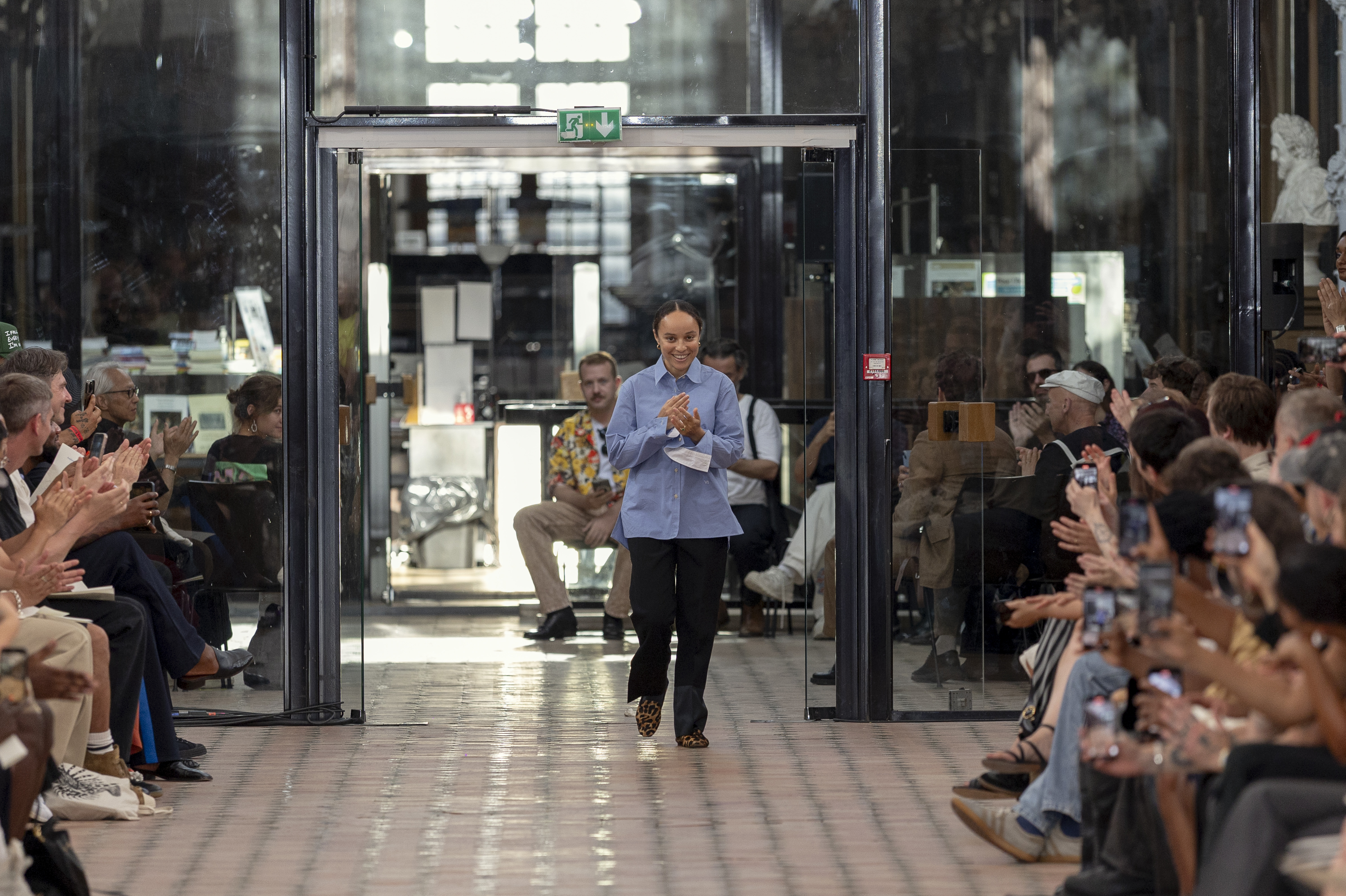 A fashion show runway with a designer walking down the aisle, applauded by the audience. The setting is modern and stylish.
