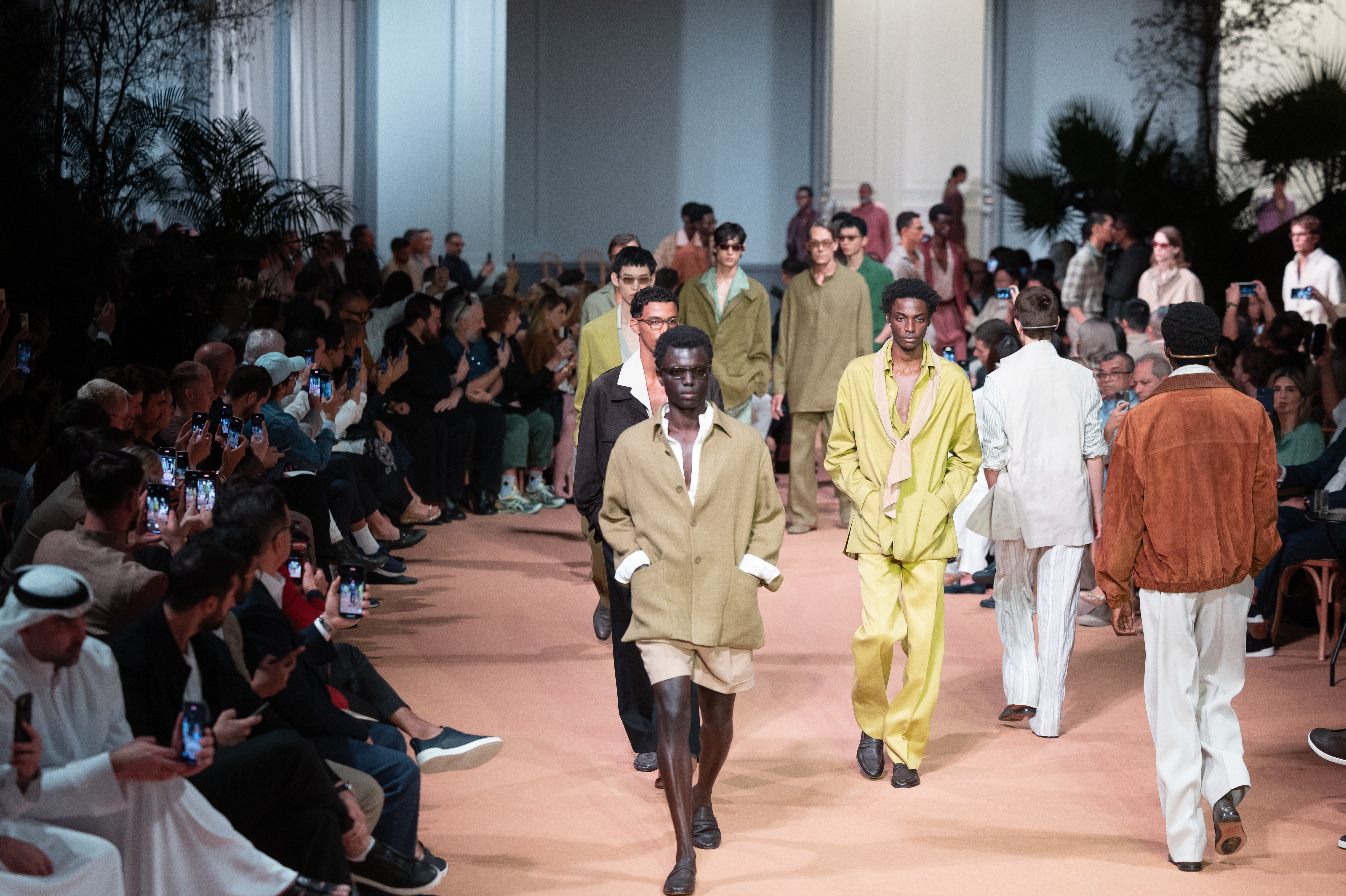 Models walking on a fashion runway, wearing earthy-toned outfits. The audience is seated on either side, capturing the show on their phones.