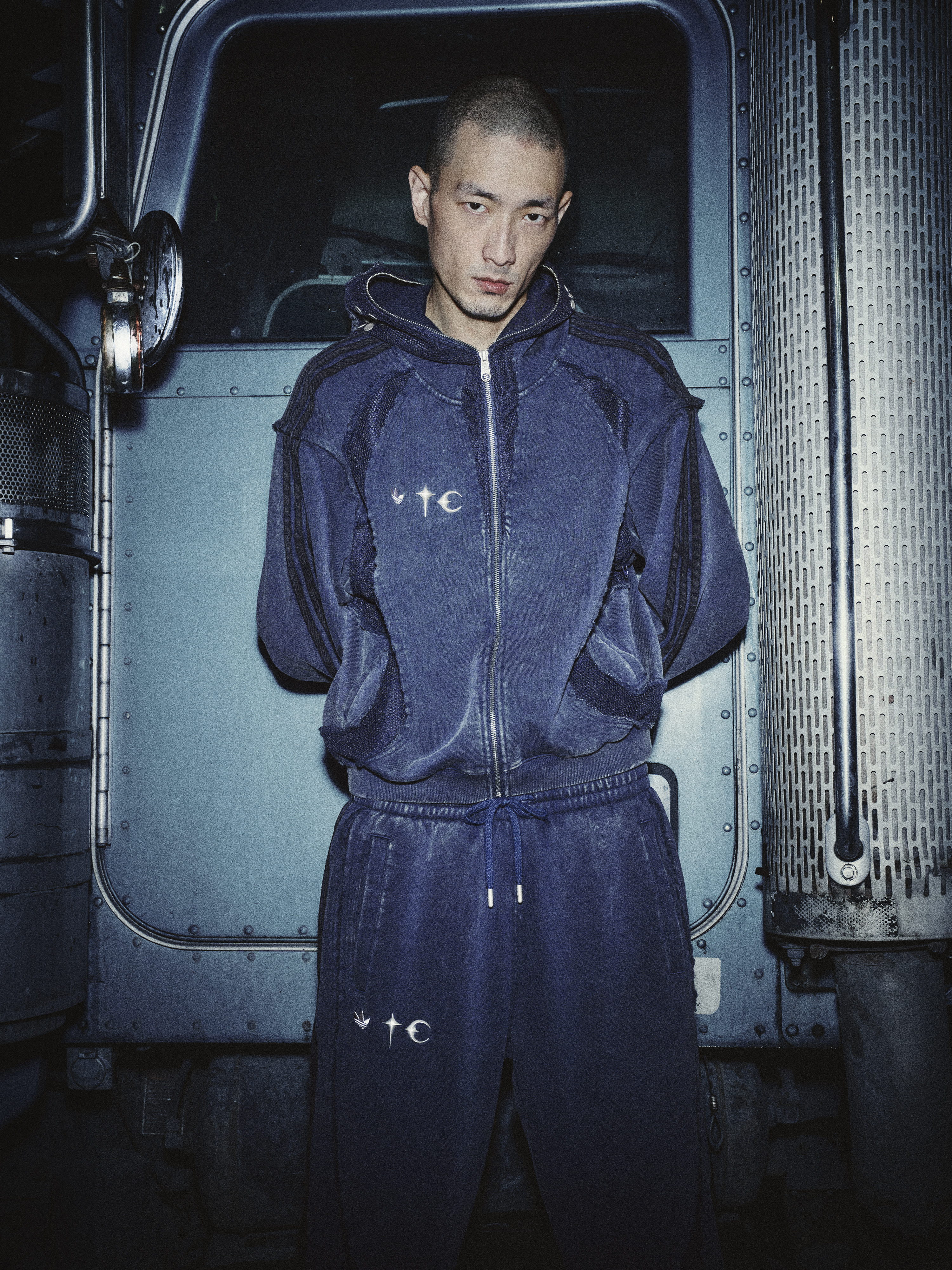 A person in a blue tracksuit stands in front of a truck, looking directly at the camera.