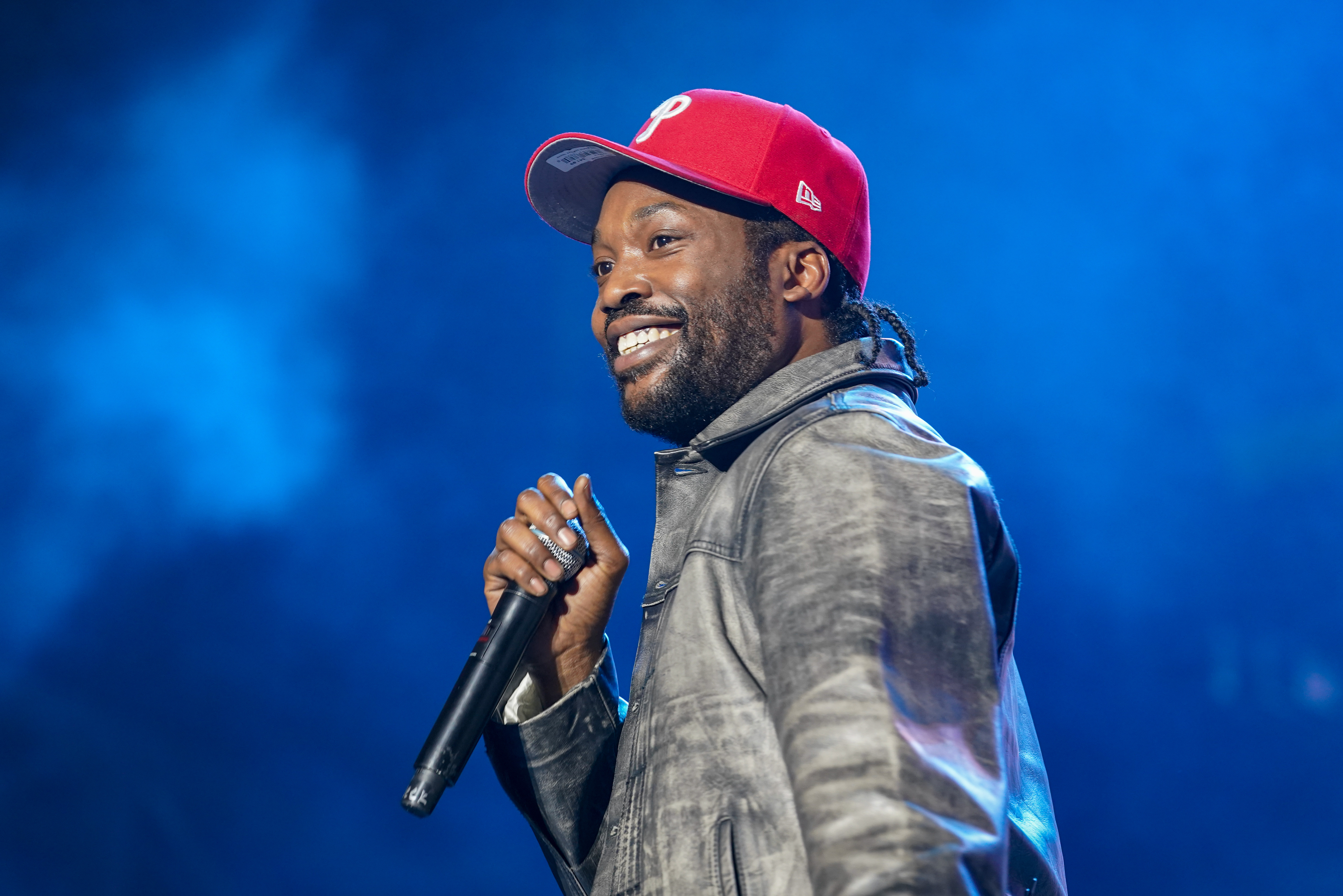 Meek Mill in a red cap and leather jacket smiles while holding a microphone on stage, with a blue background.