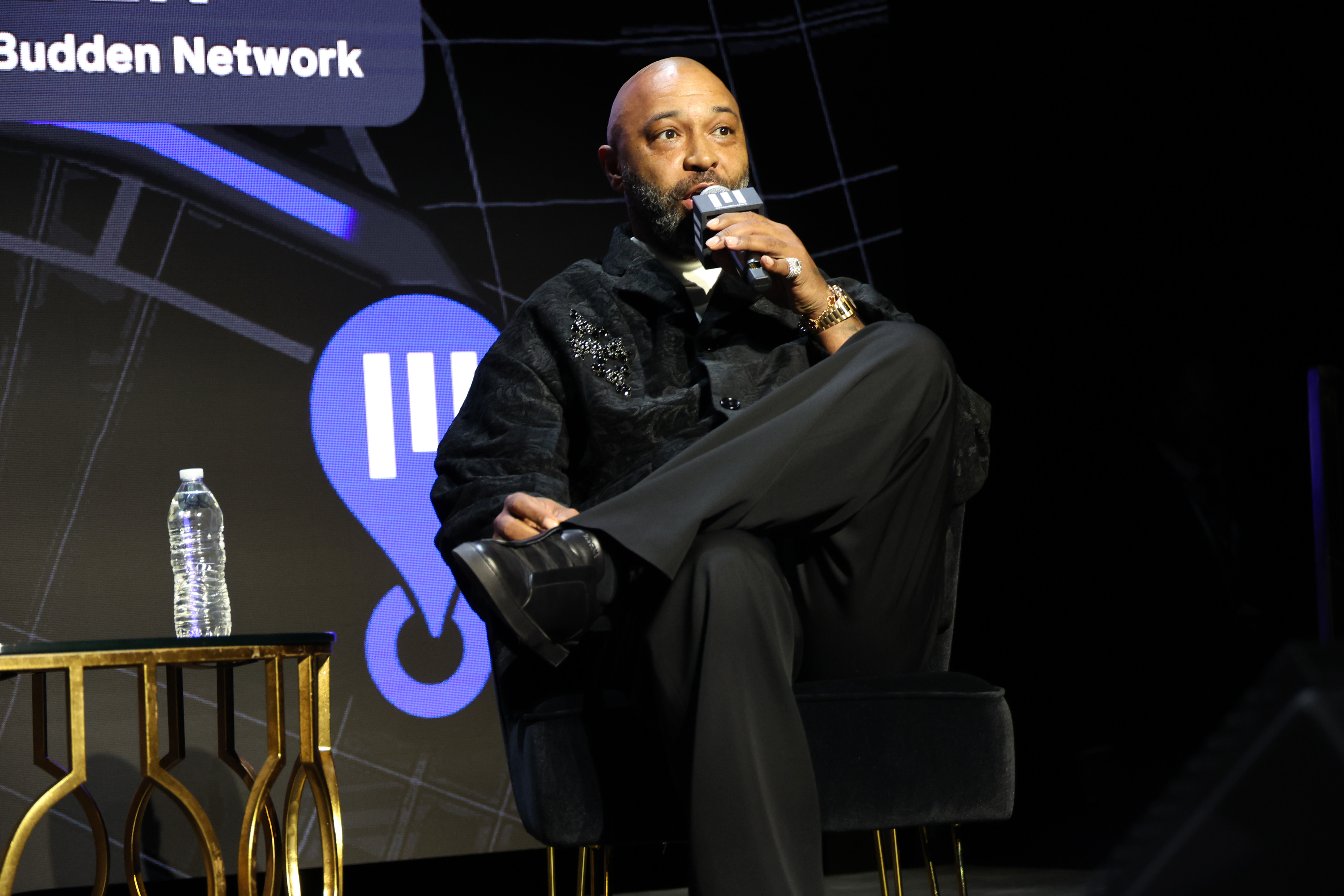 Joe Budden on stage holding a microphone, wearing a dark outfit. A table with a water bottle is beside him. Event backdrop visible.