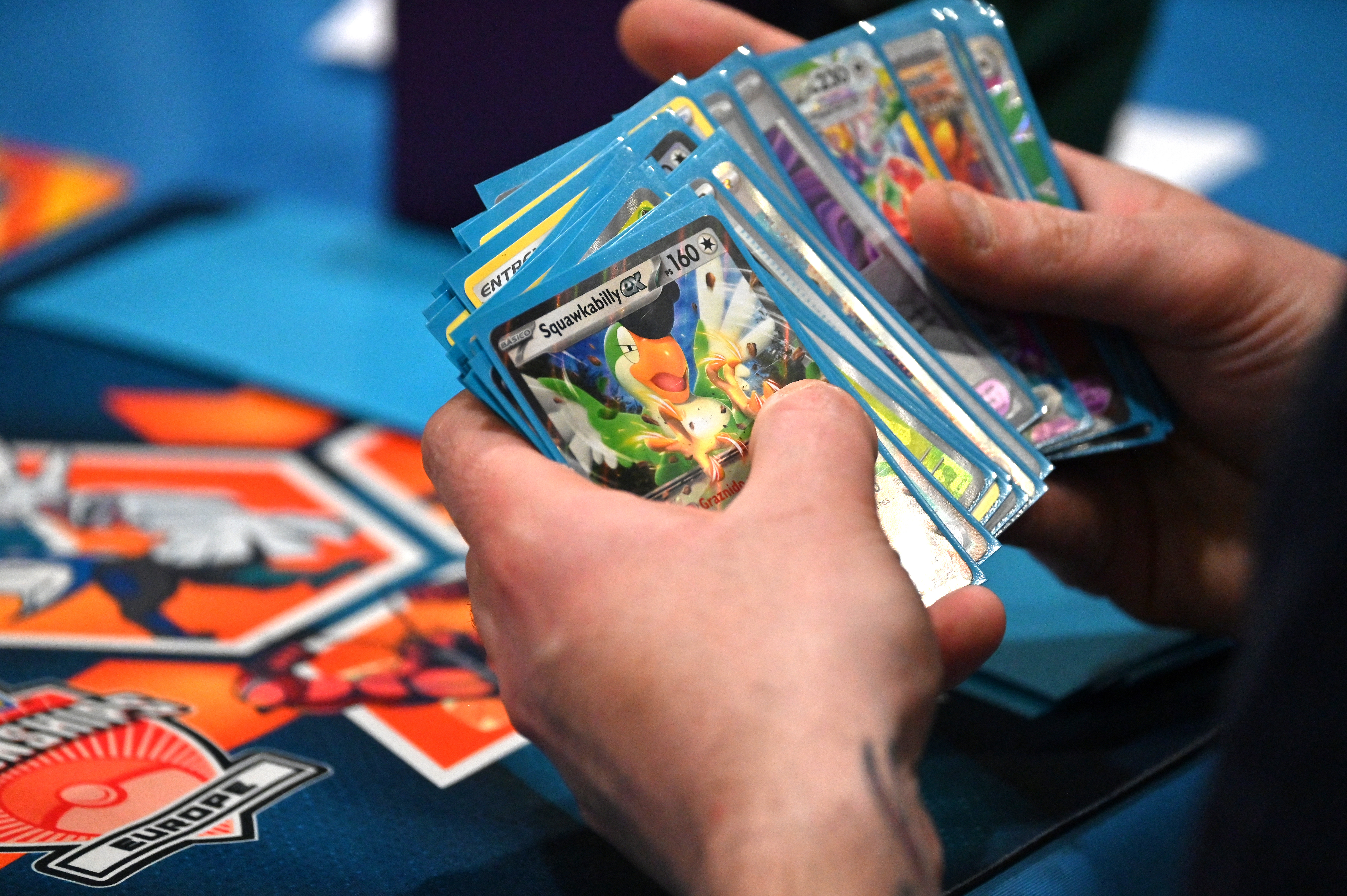 A person holds a deck of Pokémon cards, including a Squawkabilly card, over a colorful game mat.