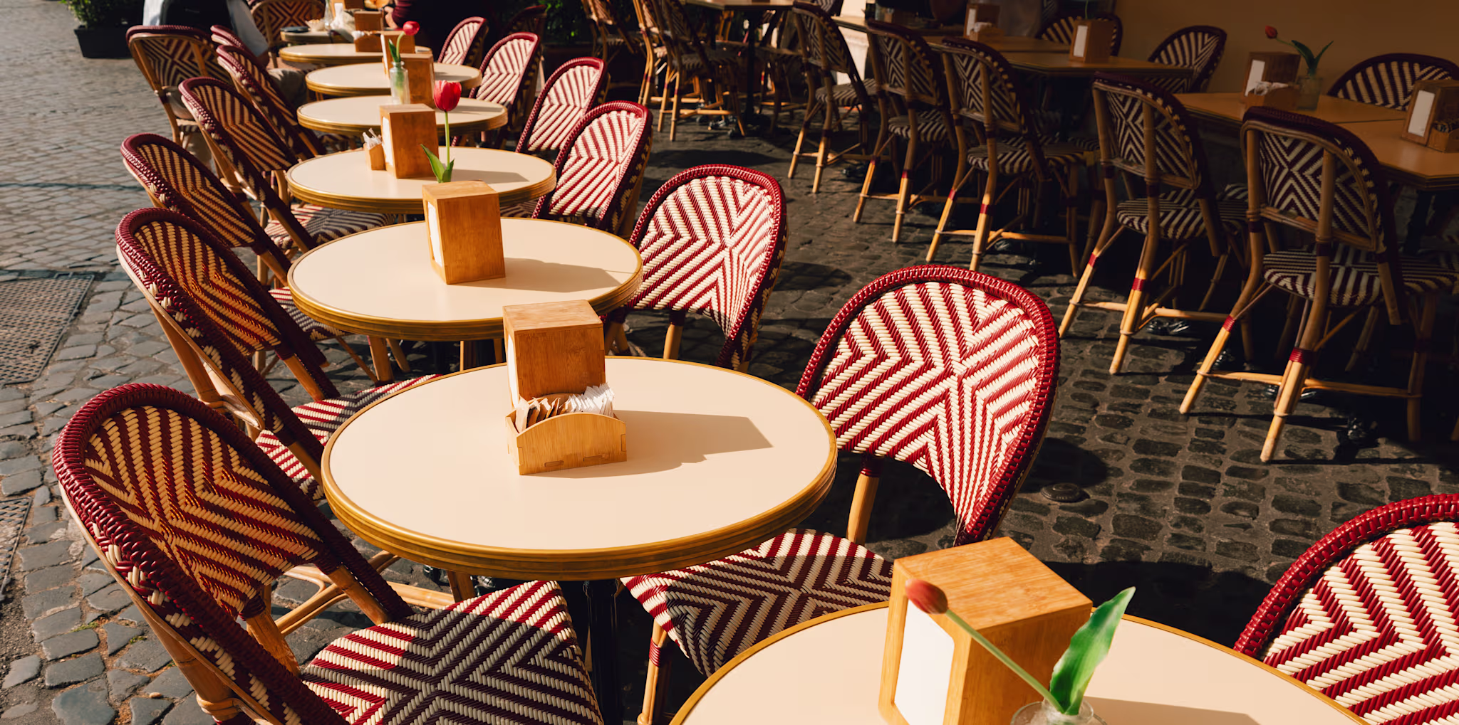 Outdoor café with round tables, wicker chairs, and cobblestone pavement. Each table has a wooden napkin holder and a small plant.