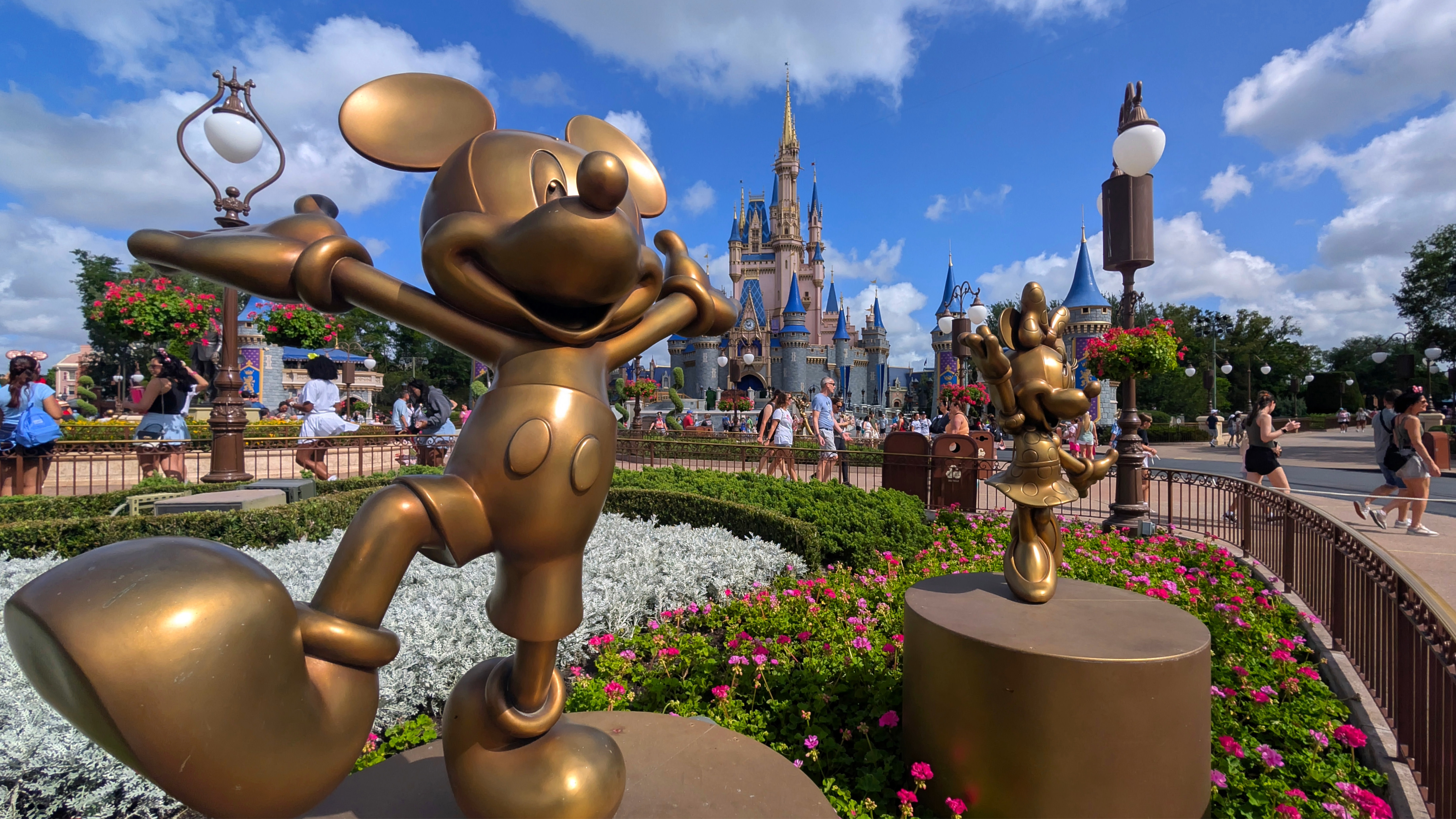 Bronze statues of Mickey and Minnie Mouse in front of Cinderella Castle at a theme park, surrounded by flowers and visitors.
