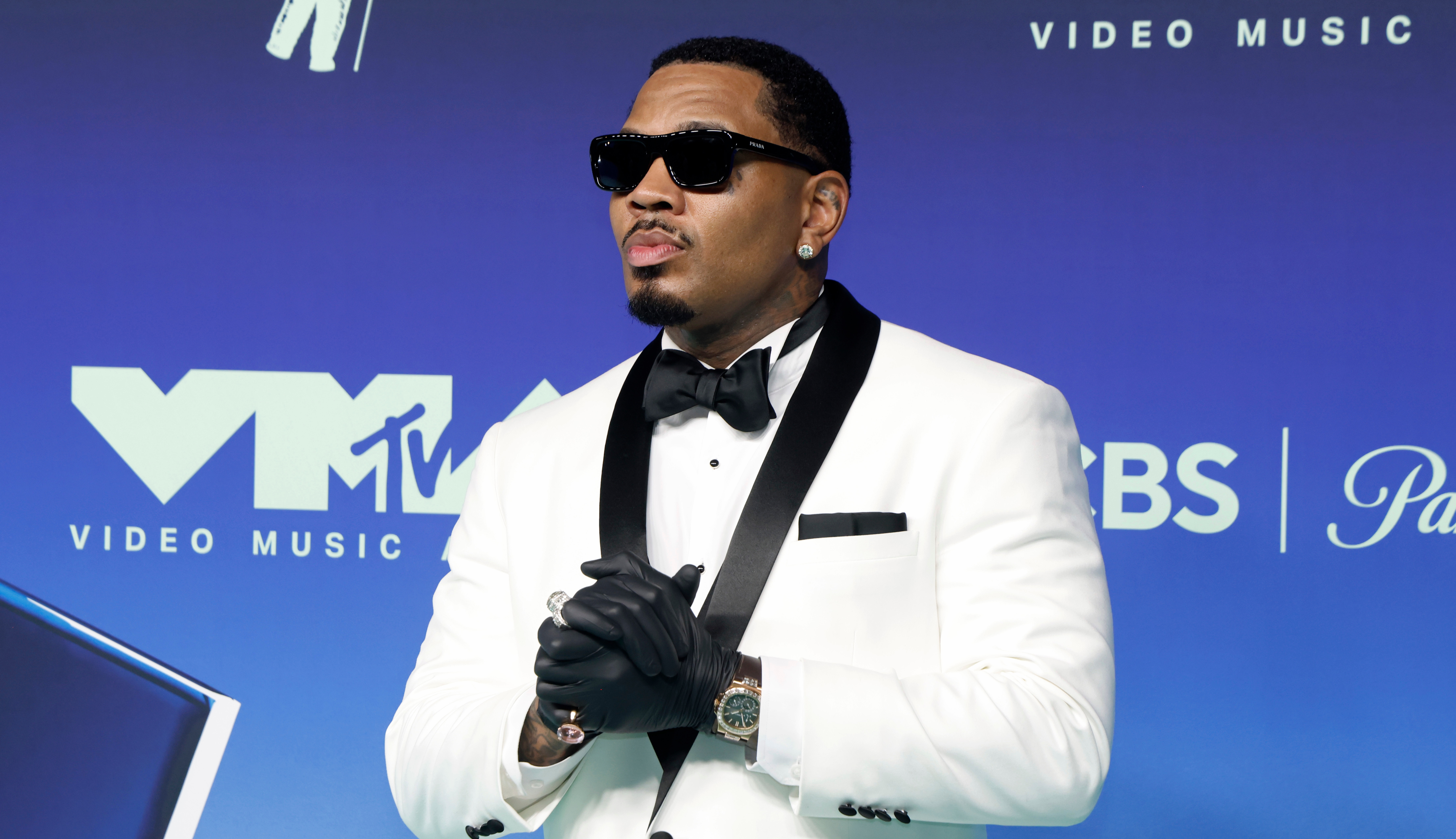 Kevin Gates in a white tuxedo with black gloves and sunglasses poses on the MTV Video Music Awards red carpet.