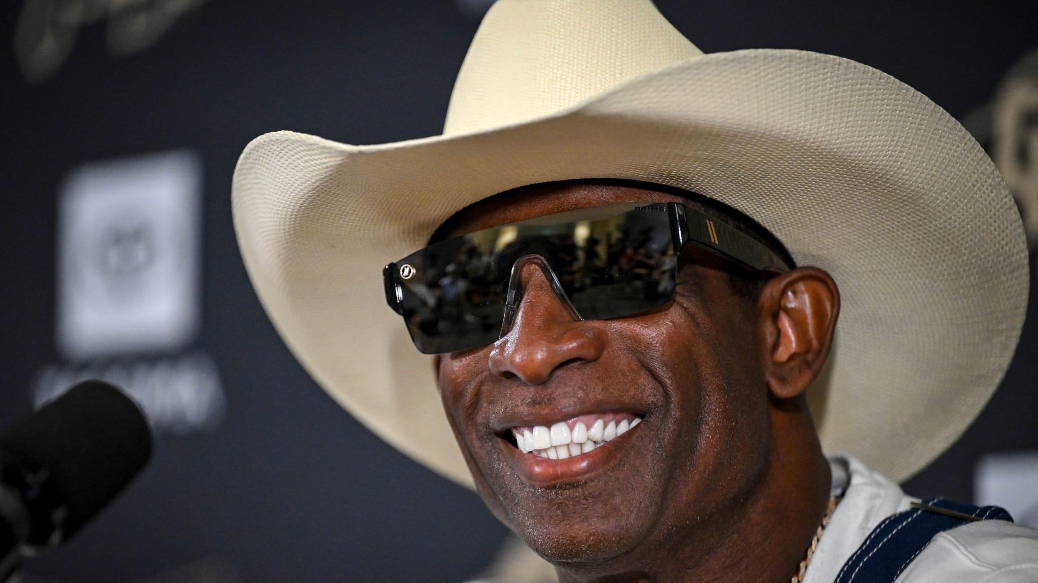 Deion Sanders wearing a cowboy hat and sunglasses, smiling at a press event.
