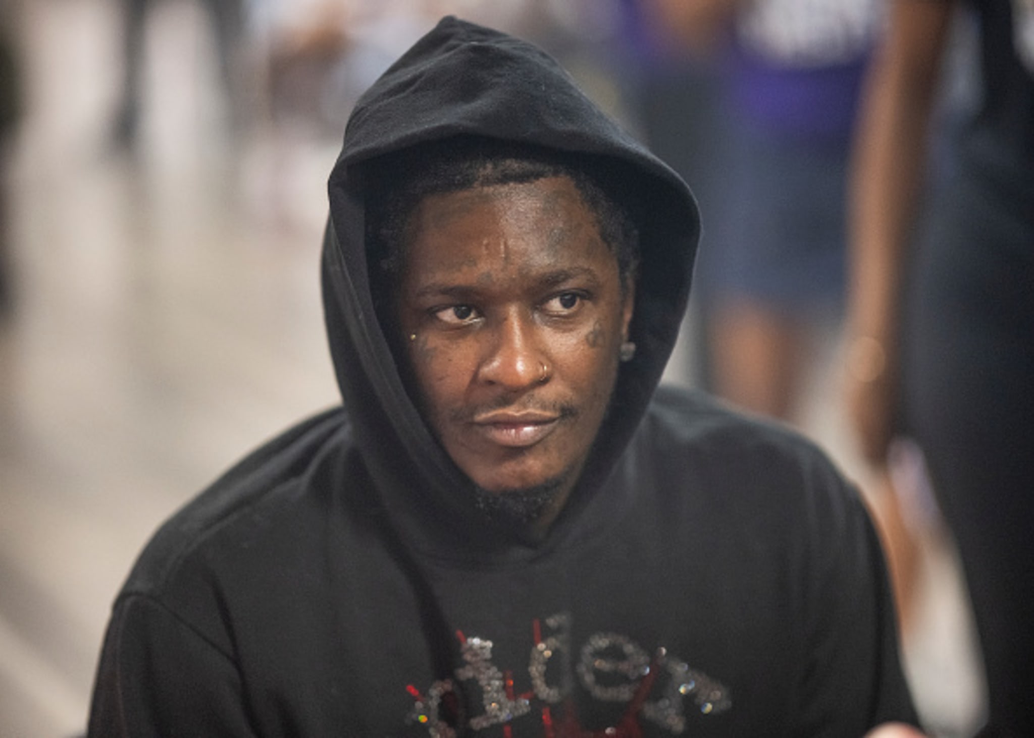 A person wearing a black hoodie with a pensive expression in a blurred background setting.