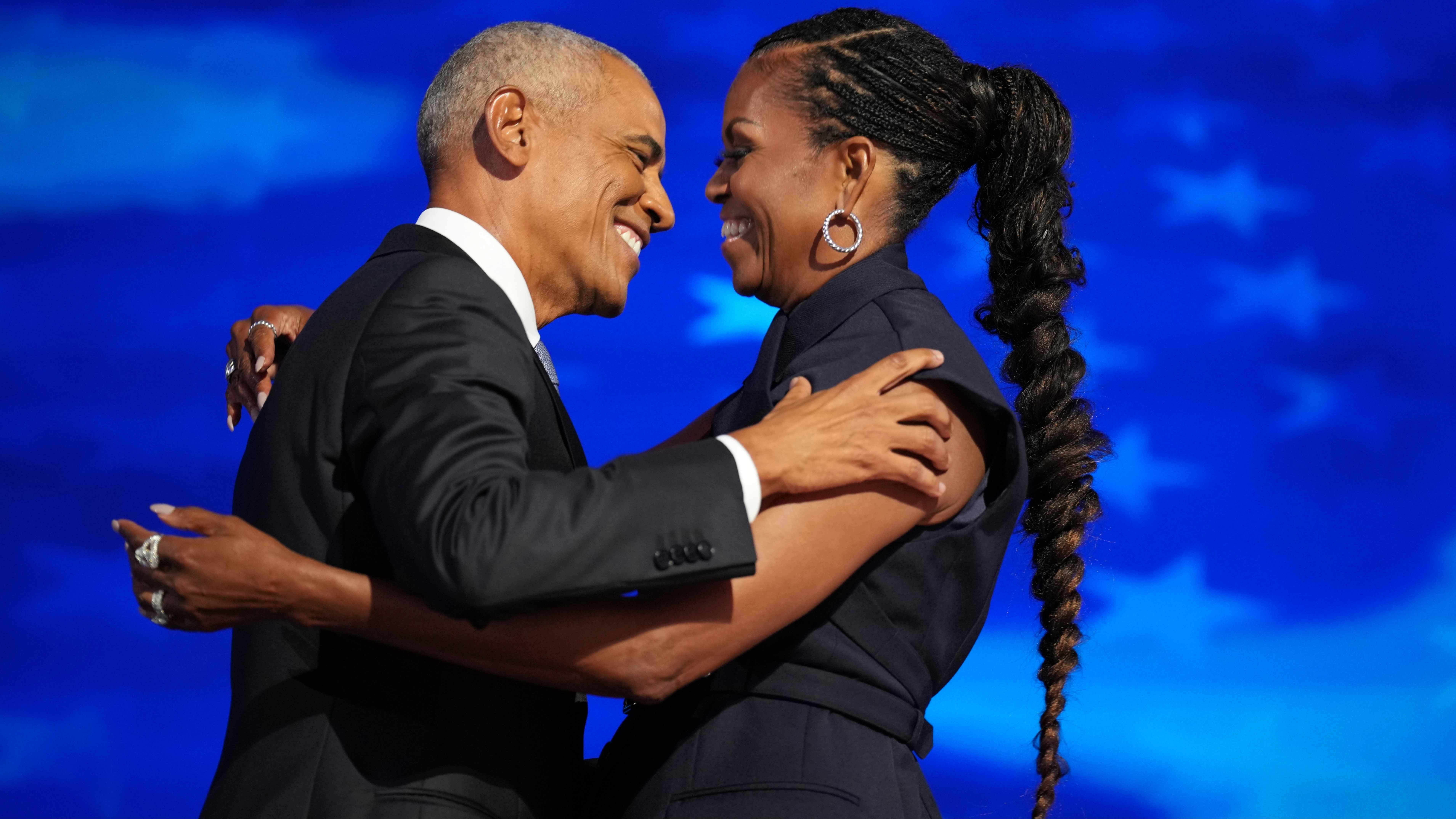 Barack Obama and Michelle Obama embrace on stage, smiling warmly at each other against a blue background with star patterns.