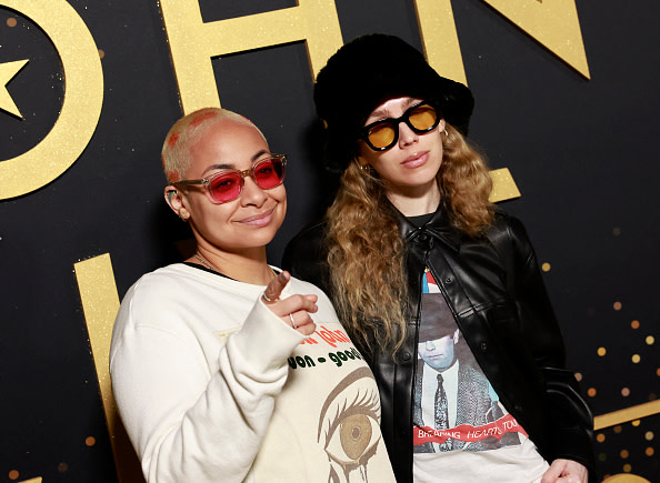 Raven-Symoné, with short hair and red glasses, stands next to a person in a black hat and sunglasses at an event.