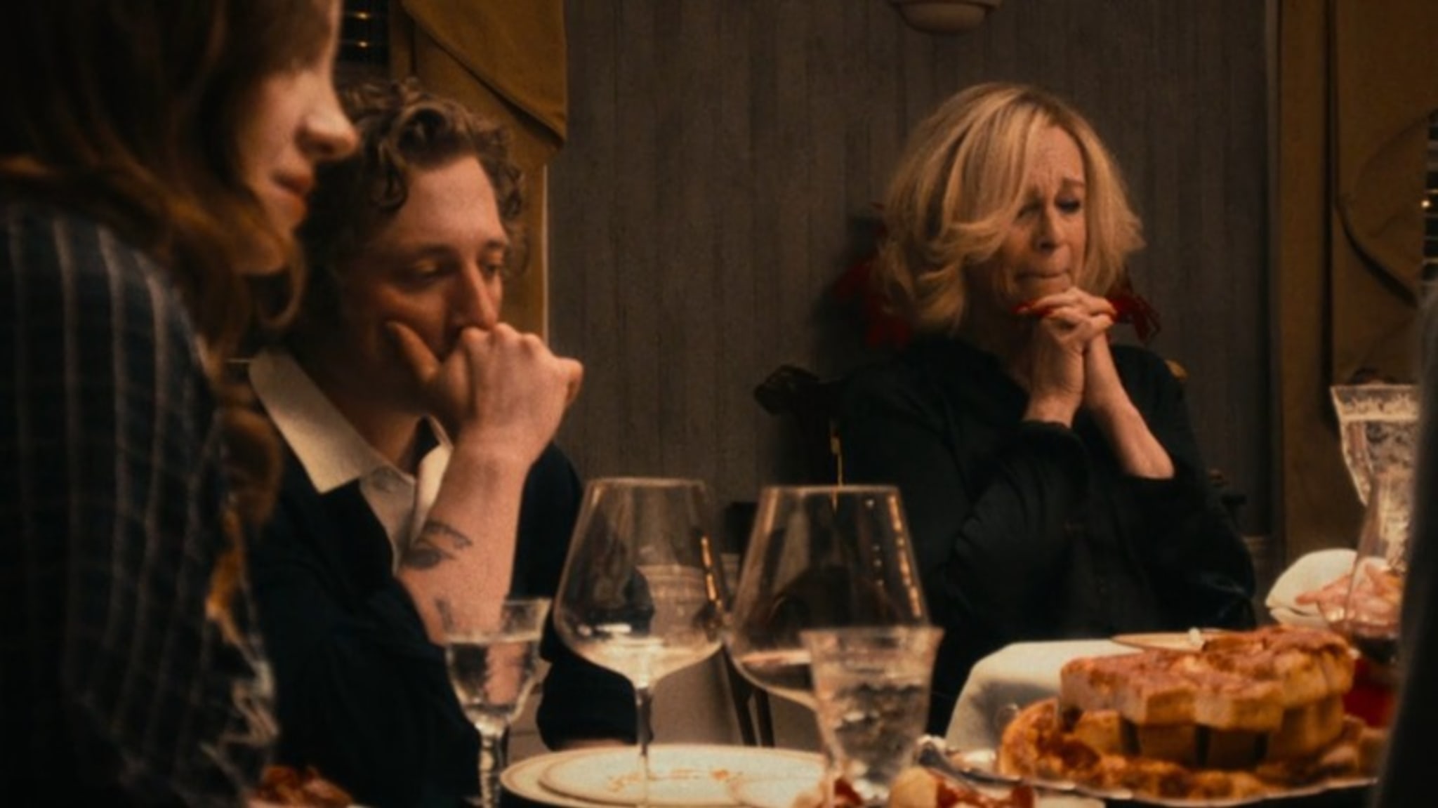 A group of people sitting at a dinner table, appearing thoughtful or contemplative, with food and glasses in front of them.