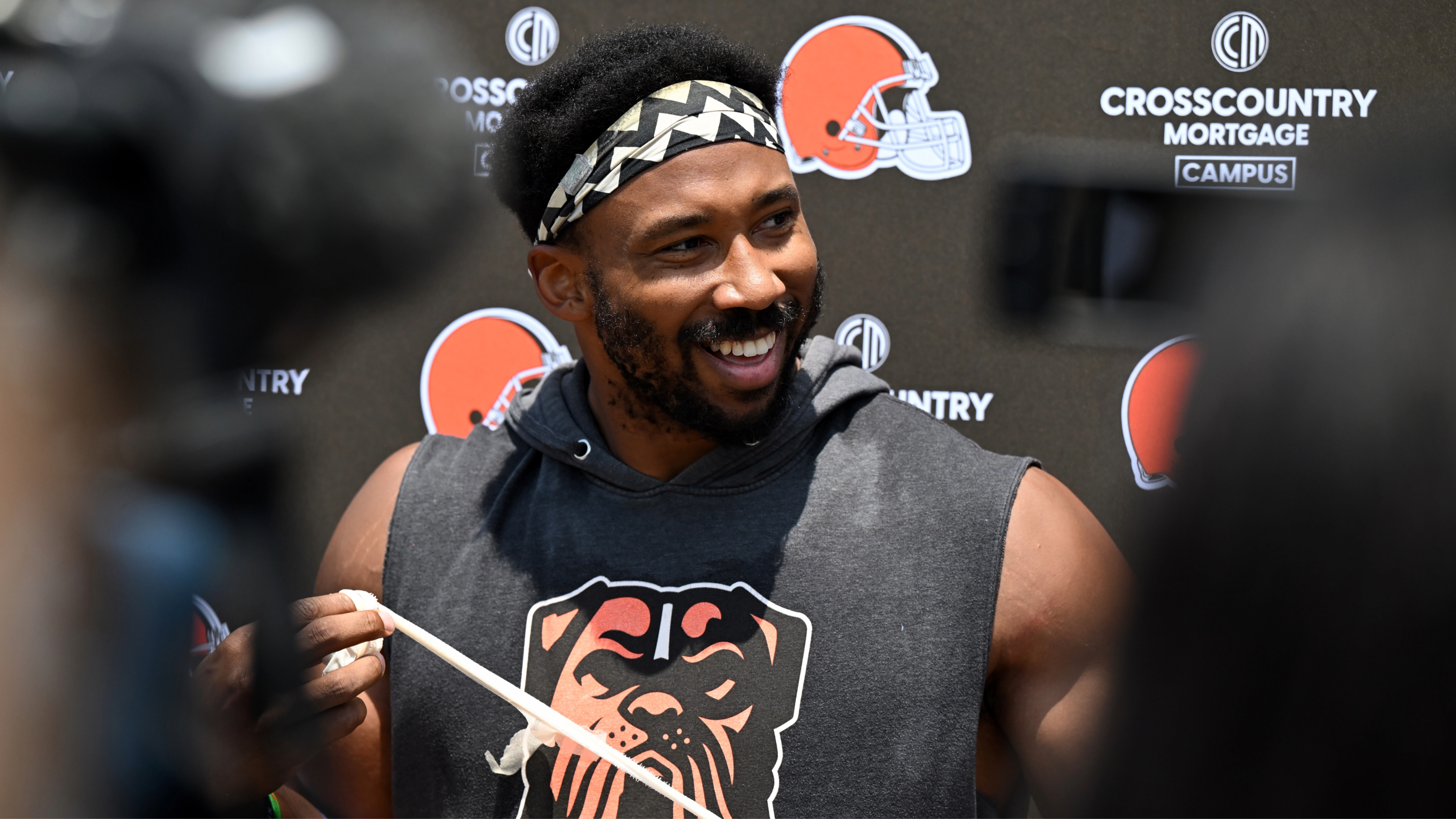 Myles Garrett with a headband and sleeveless hoodie smiles at a press event, with Cleveland Browns logos in the background.