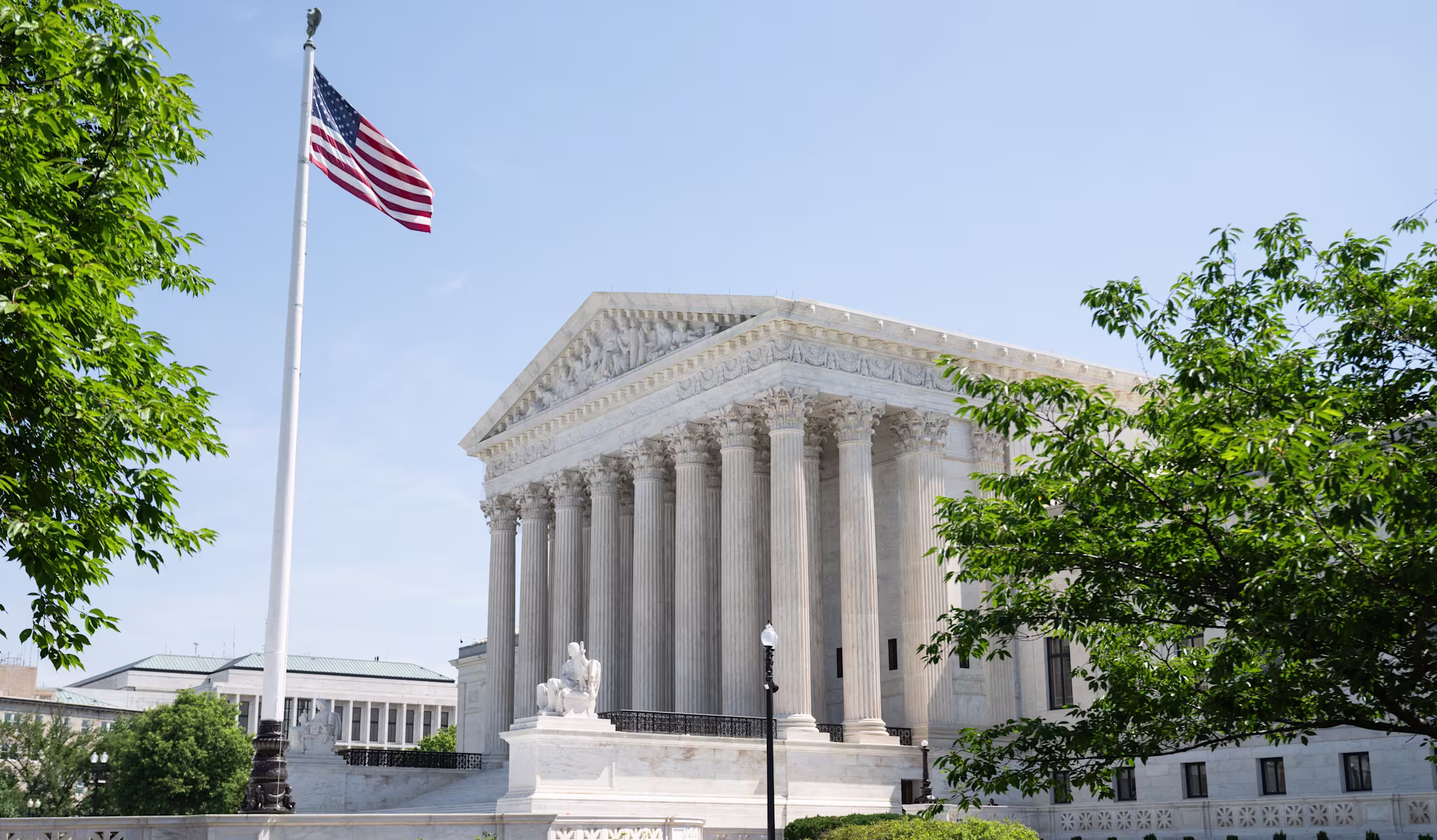 The image shows the United States Supreme Court building with columns, an American flag, and surrounding greenery under a clear blue sky.