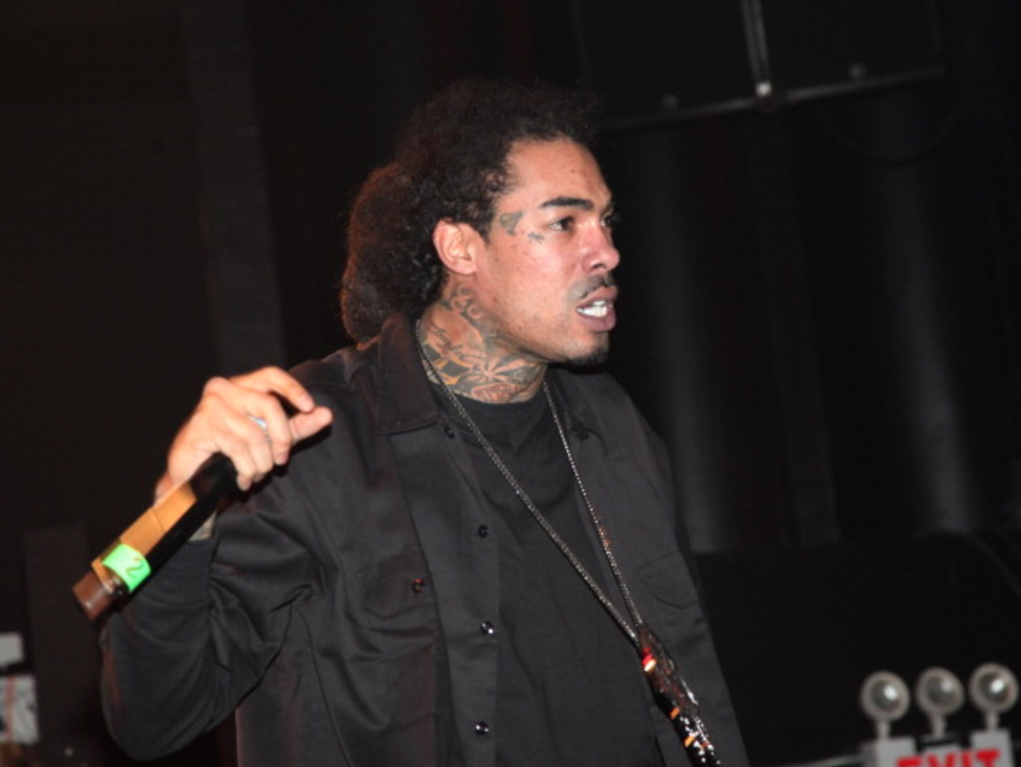 Gunplay performing on stage, holding a microphone, wearing a black outfit.