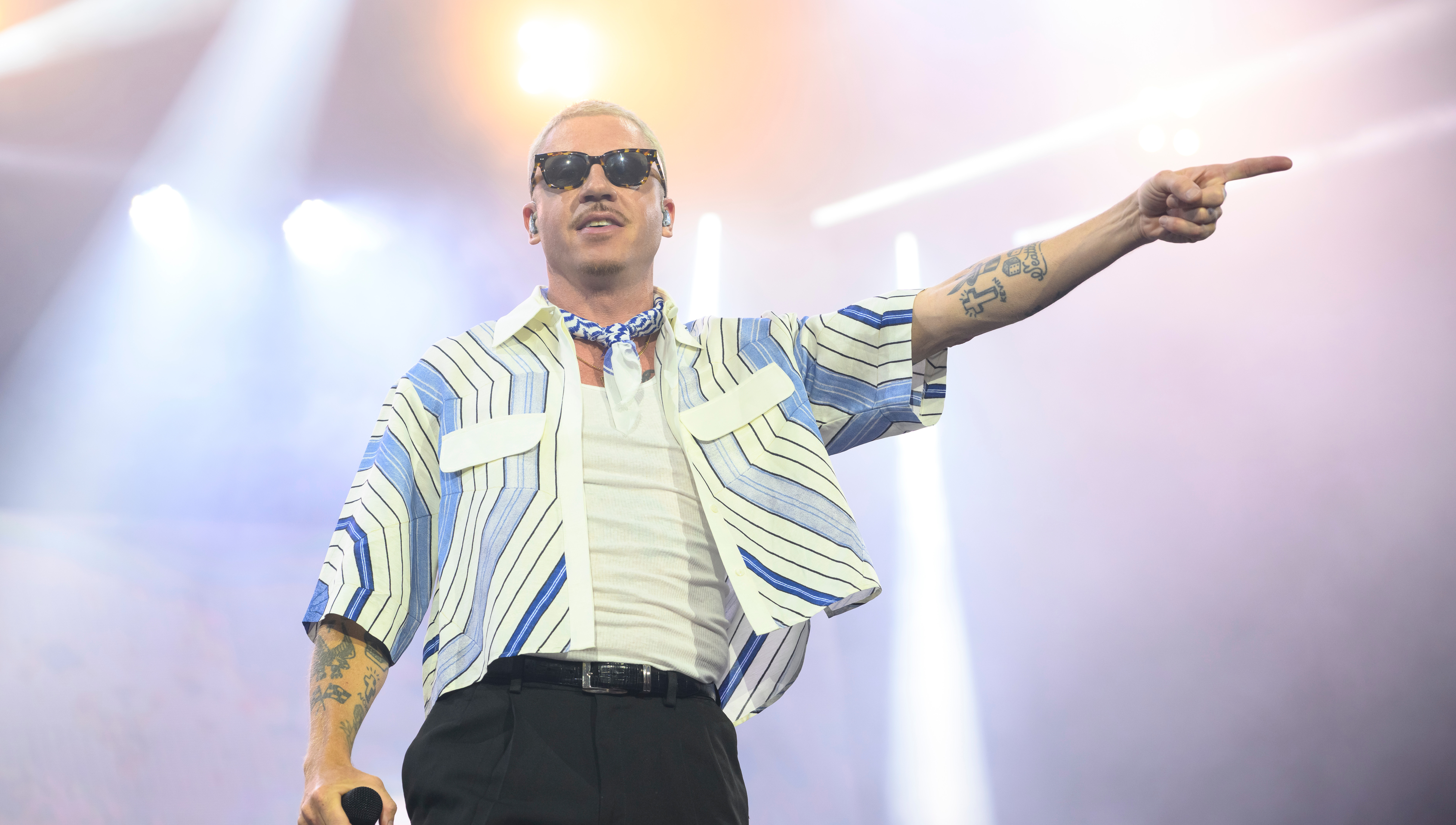 Macklemore wearing sunglasses and a striped shirt points while holding a microphone on stage, with bright lights in the background.