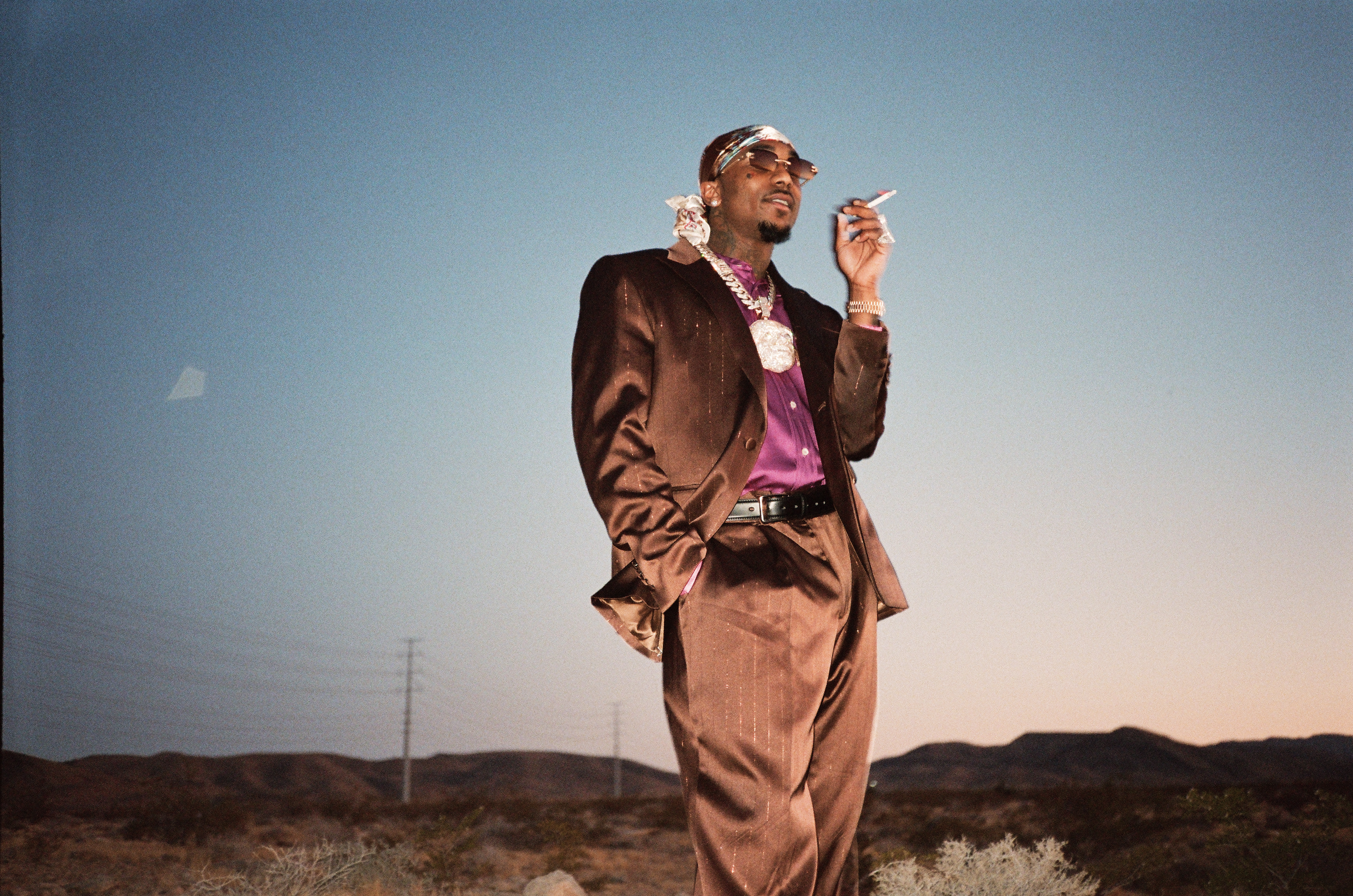 Loe Shimmy in a brown suit and purple shirt stands in a desert landscape, wearing sunglasses and a bandana, holding a cigarette.