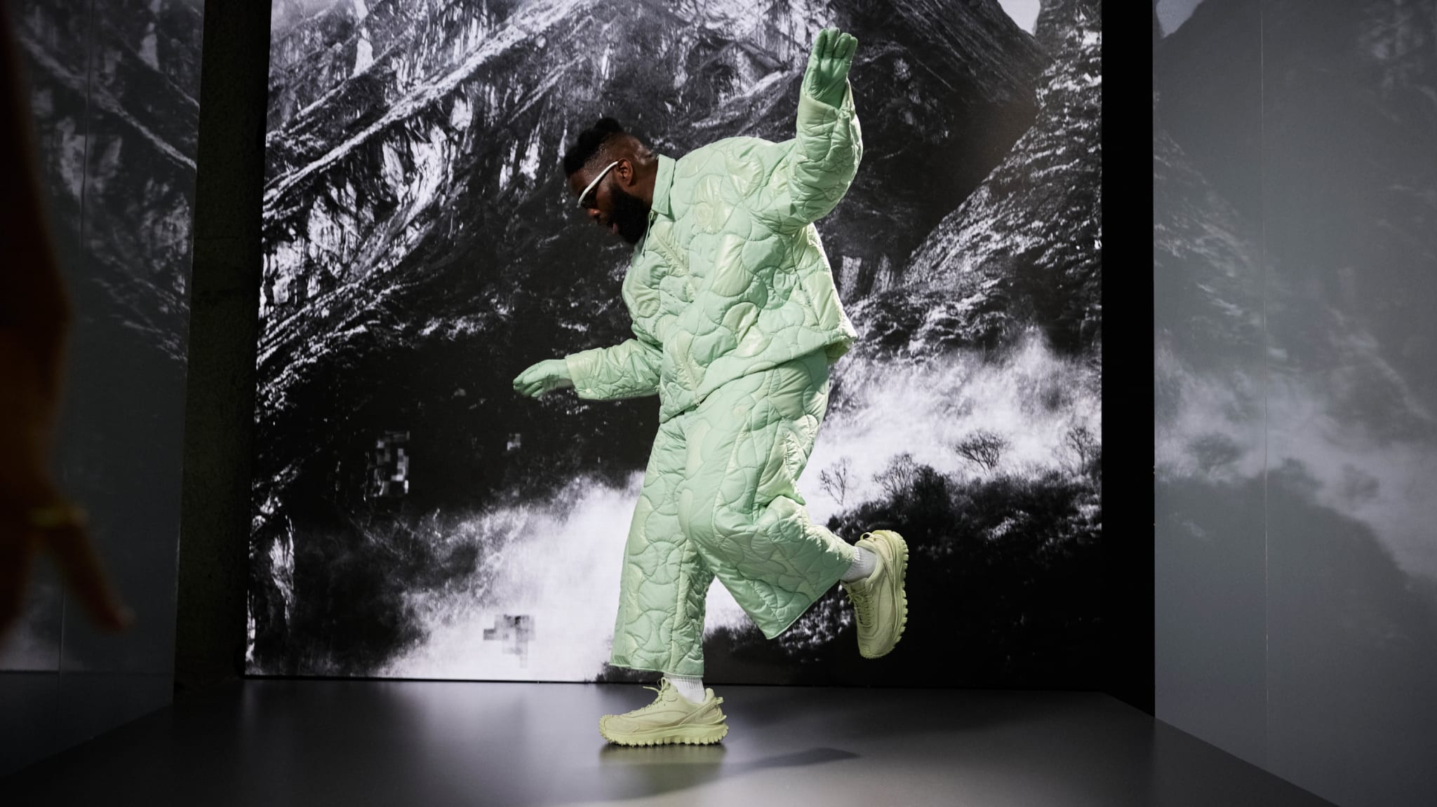 A person in a quilted light green outfit and futuristic shoes poses against a mountain backdrop.