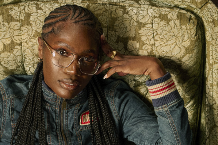 A person with braided hair and glasses sits on a patterned chair, wearing a denim jacket, plaid skirt, and orange sneakers.