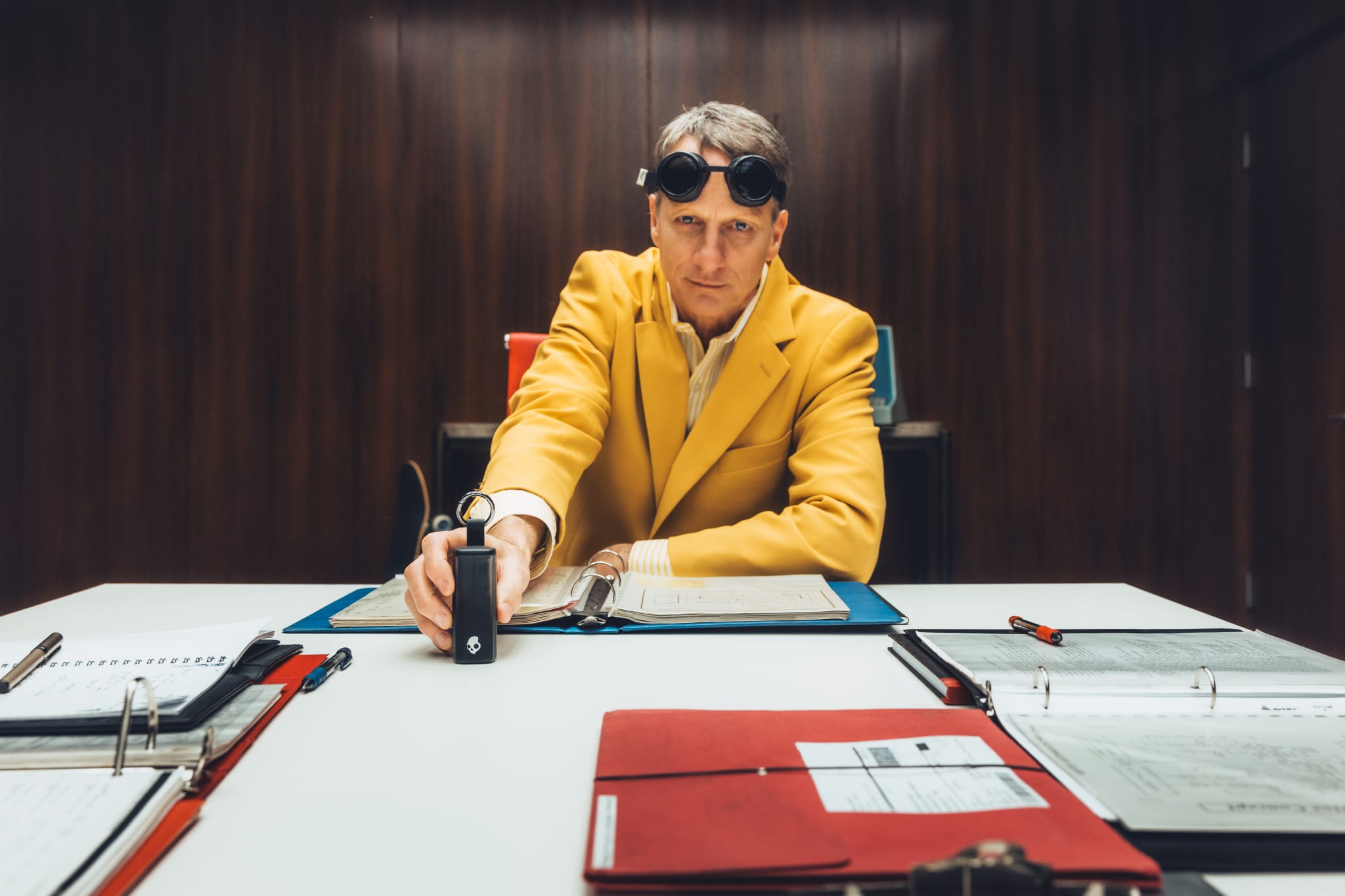 A man in a yellow suit and goggles sits at a table with binders and documents, holding a device, in a wood-paneled room.