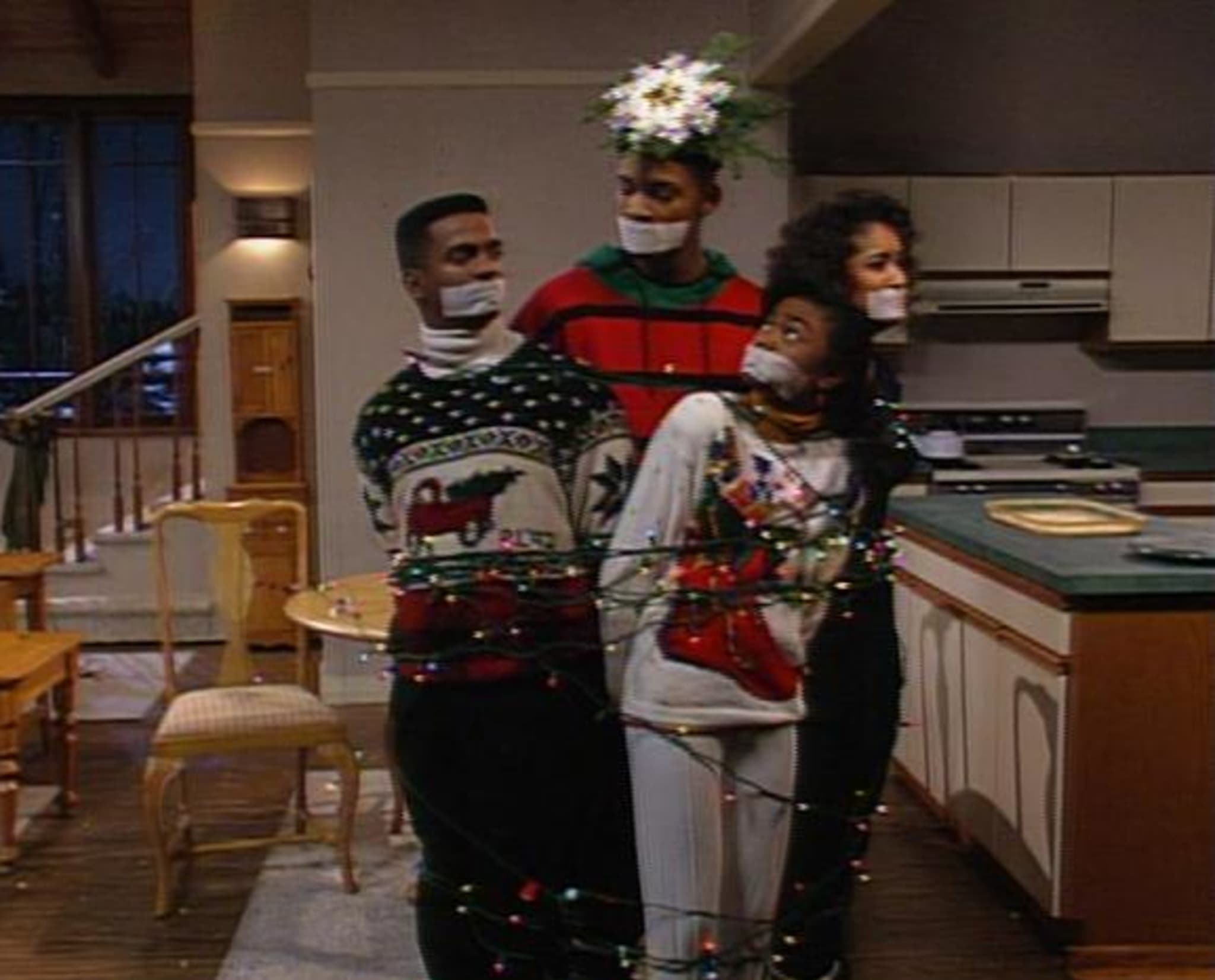 Four people in festive sweaters are tied with Christmas lights and gagged in a kitchen, with one wearing a floral headpiece.