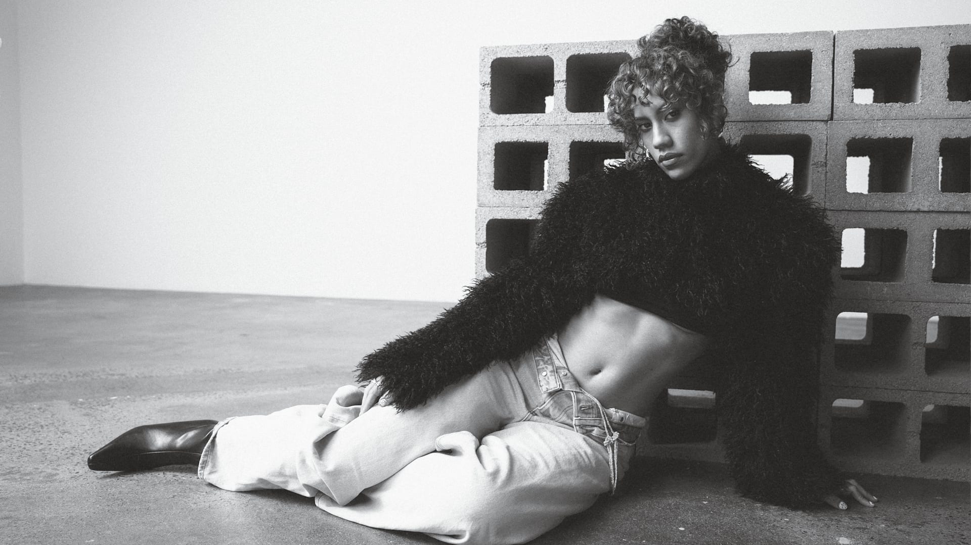A person with curly hair poses in a fuzzy sweater and jeans, seated against a stack of cinder blocks in a black and white photo.