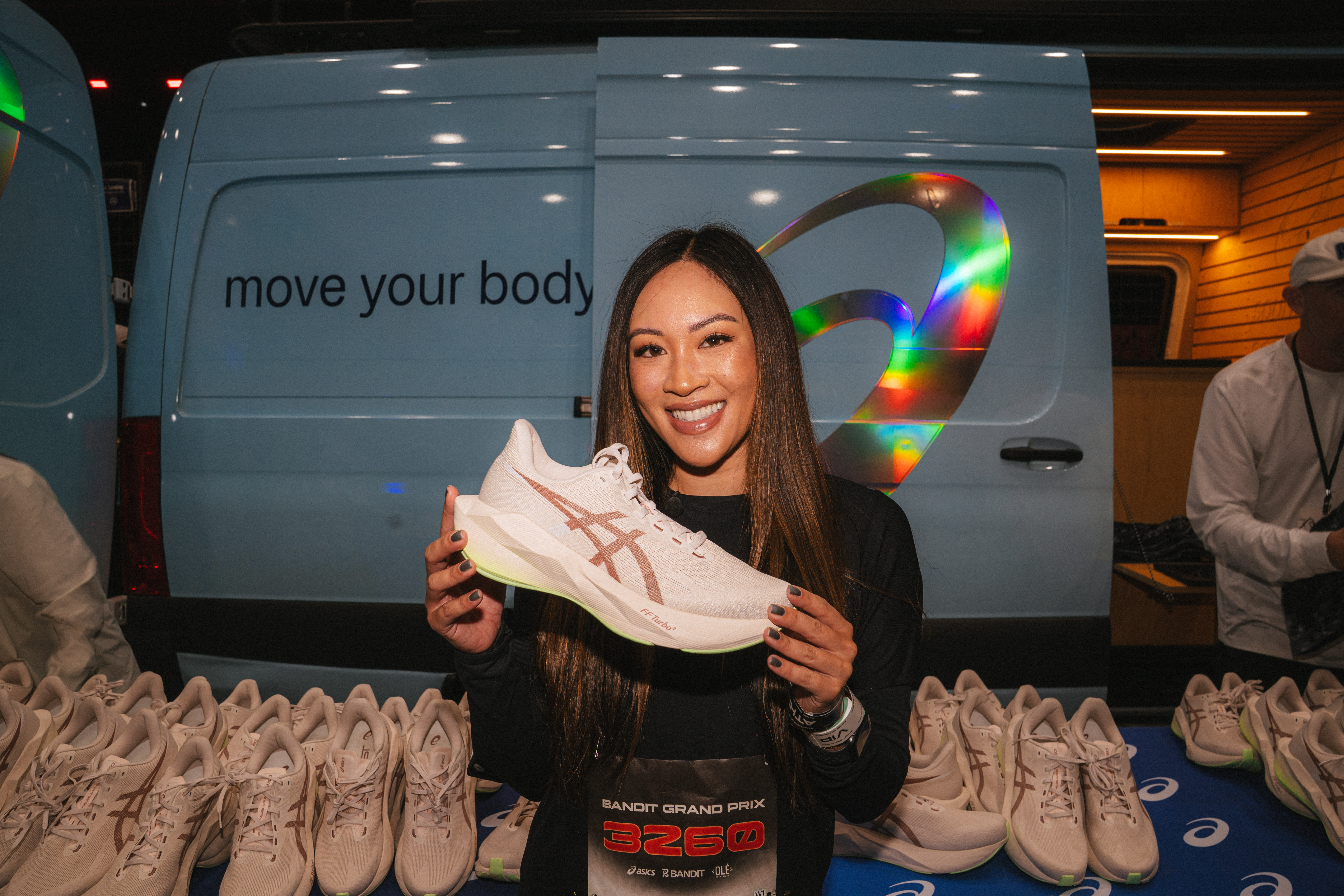 A woman smiling and holding an Asics shoe in front of a display table with more shoes. A van with "move your body" is in the background.