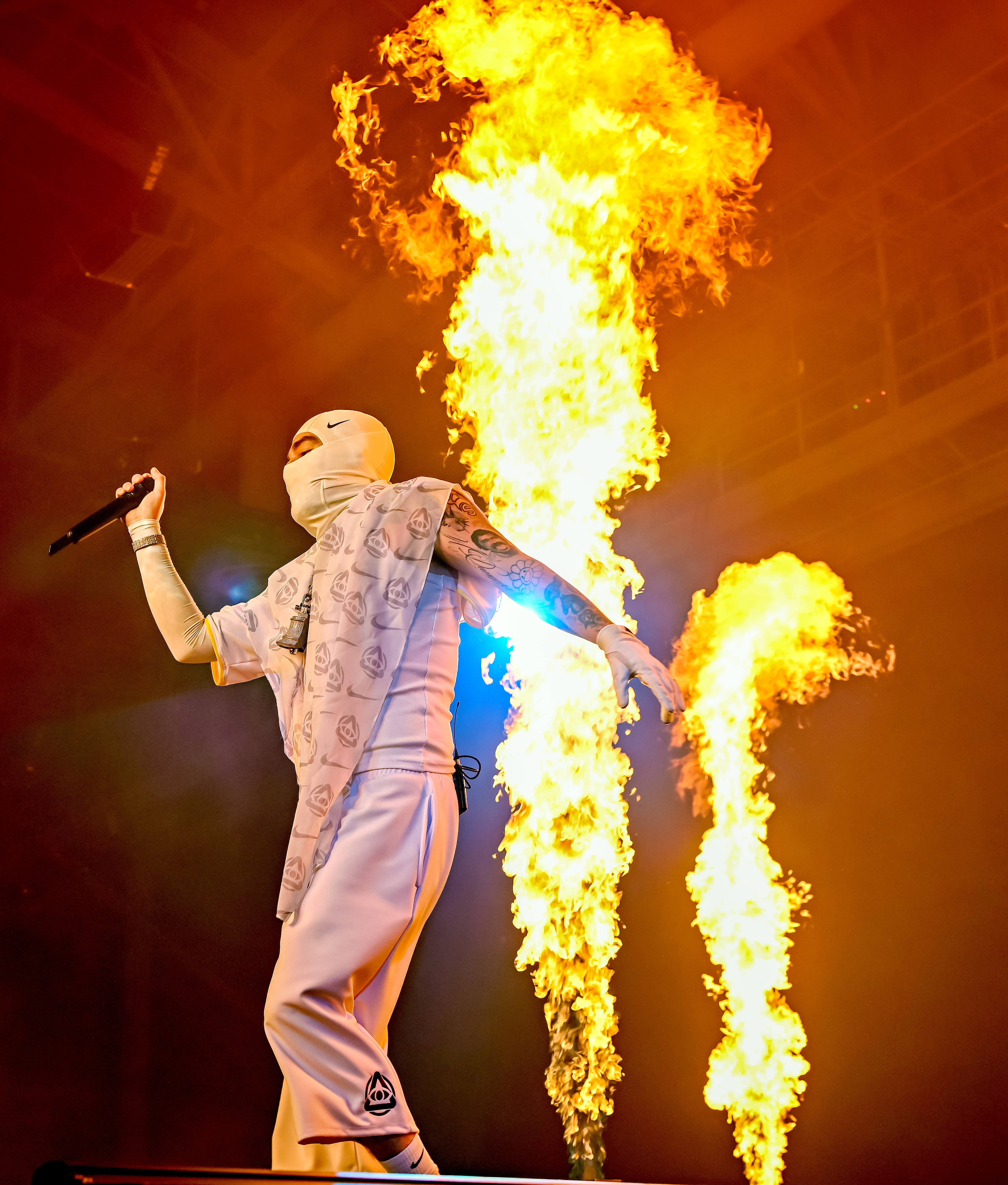 A performer wearing a white mask and outfit, holding a microphone, stands in front of large flames on stage.