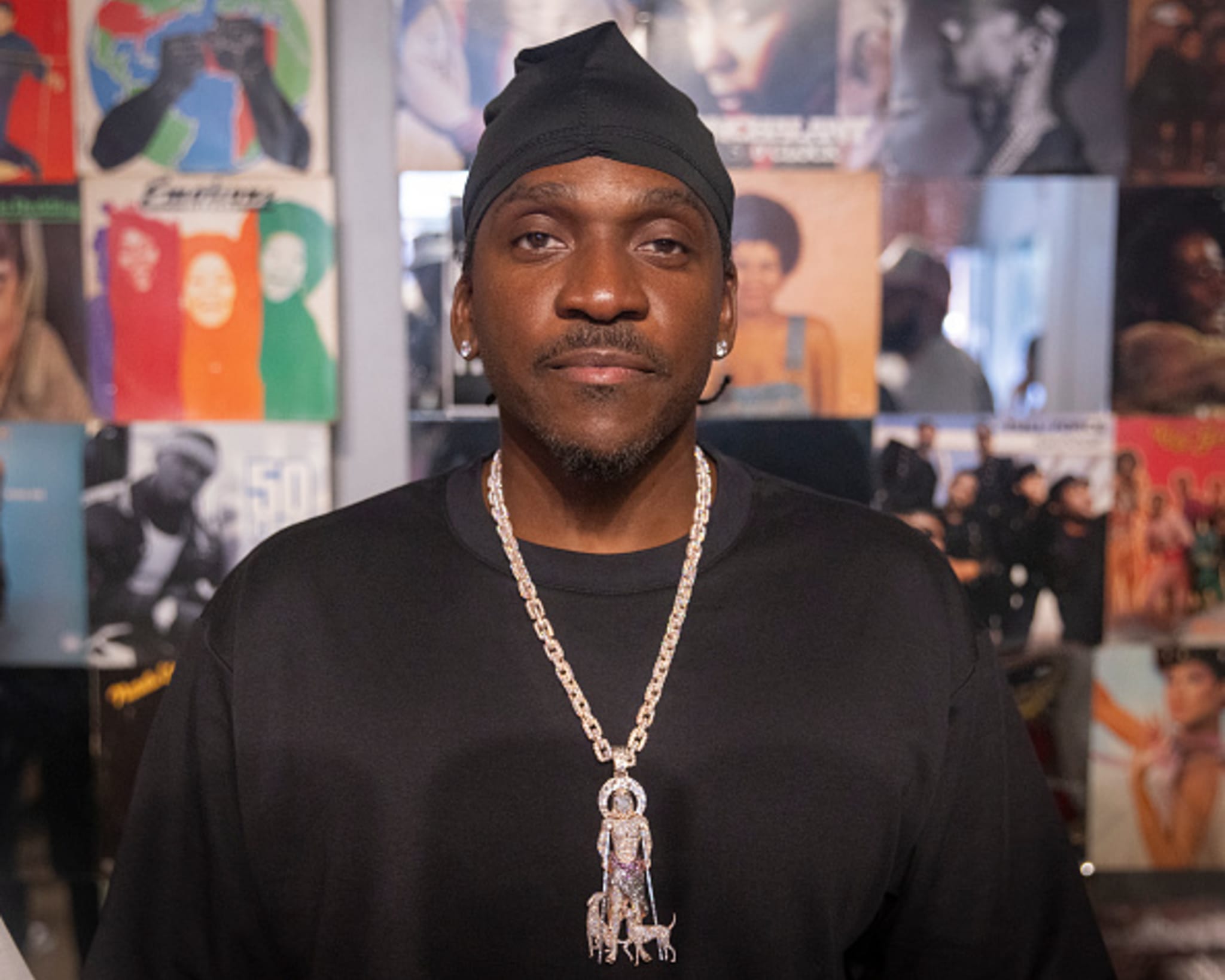 A man wearing a black shirt and a large chain necklace stands in front of a wall covered with album covers.