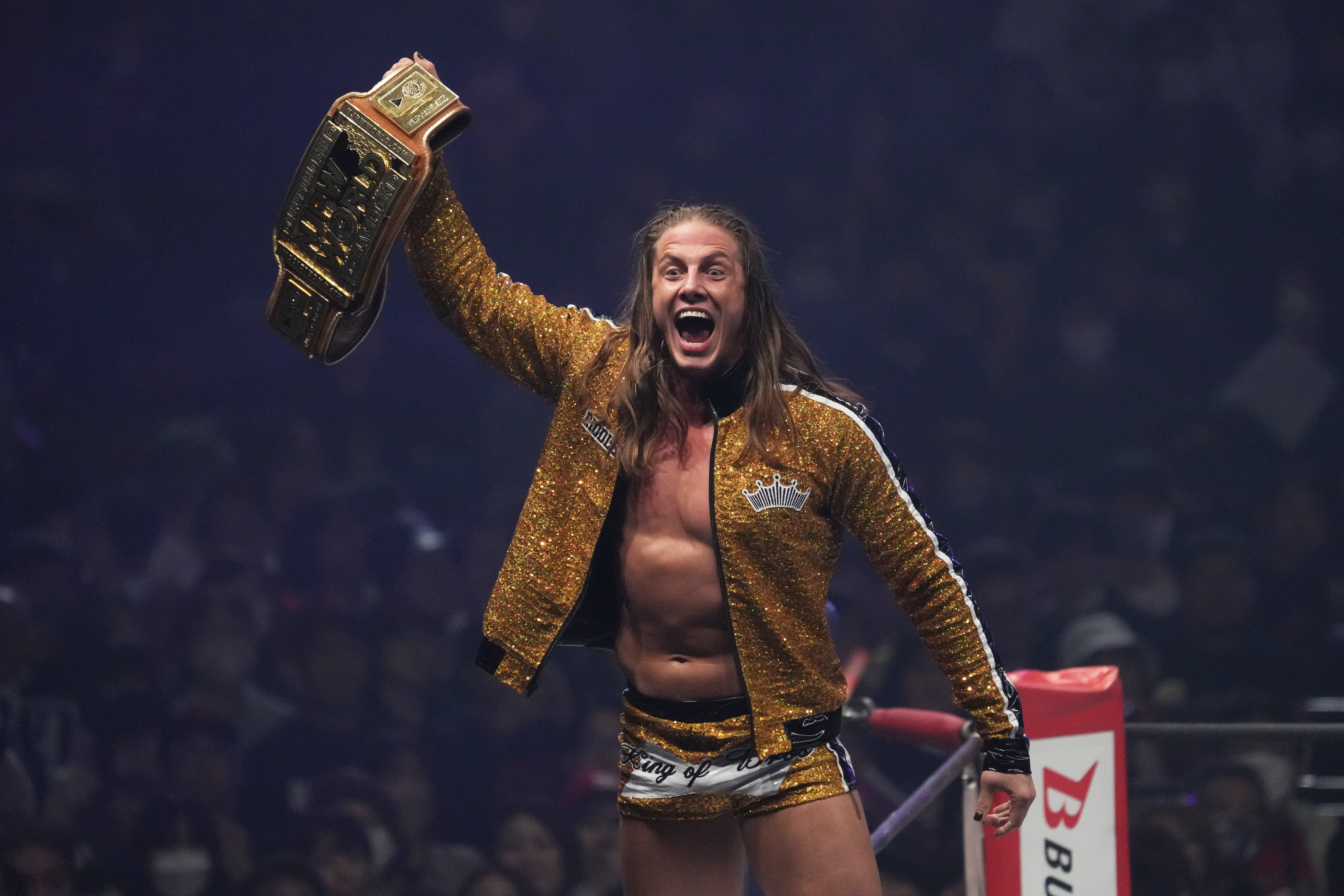 Matt Riddle with long hair and a gold jacket holds a championship belt triumphantly in a wrestling ring.