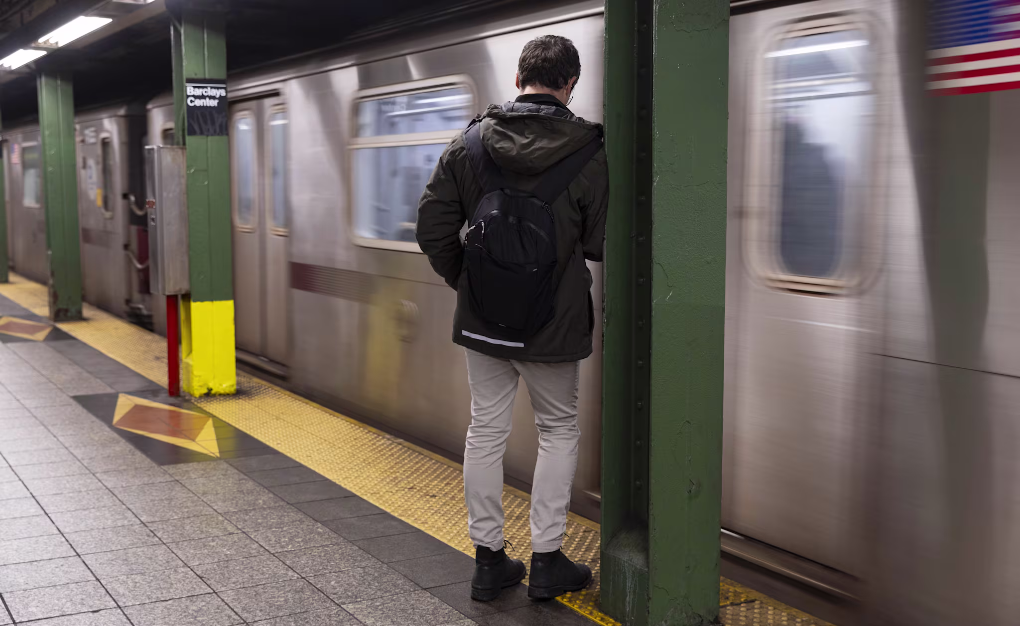 A person with a backpack waits on a subway platform as a train speeds by. Green pillars and a yellow safety line are visible.