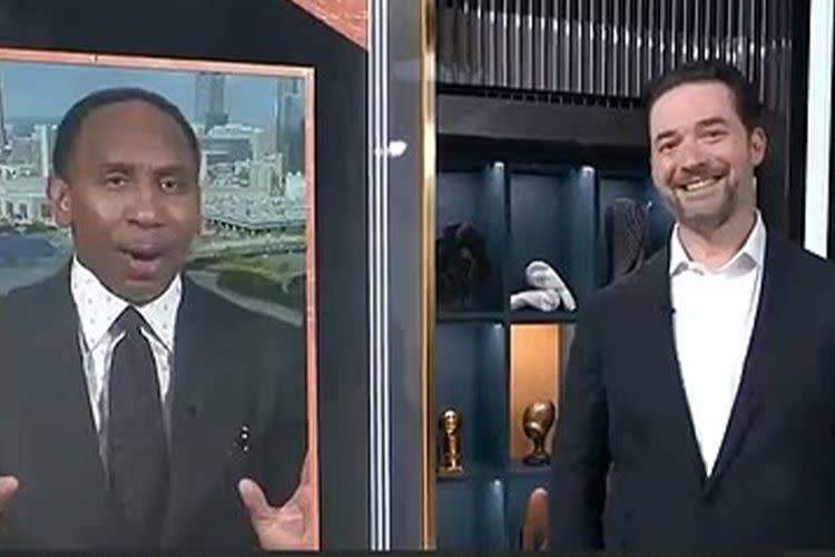A man, Stephen A. Smith, on a screen gestures animatedly, while another man, Alexis Ohanian, in a suit stands smiling beside the screen.