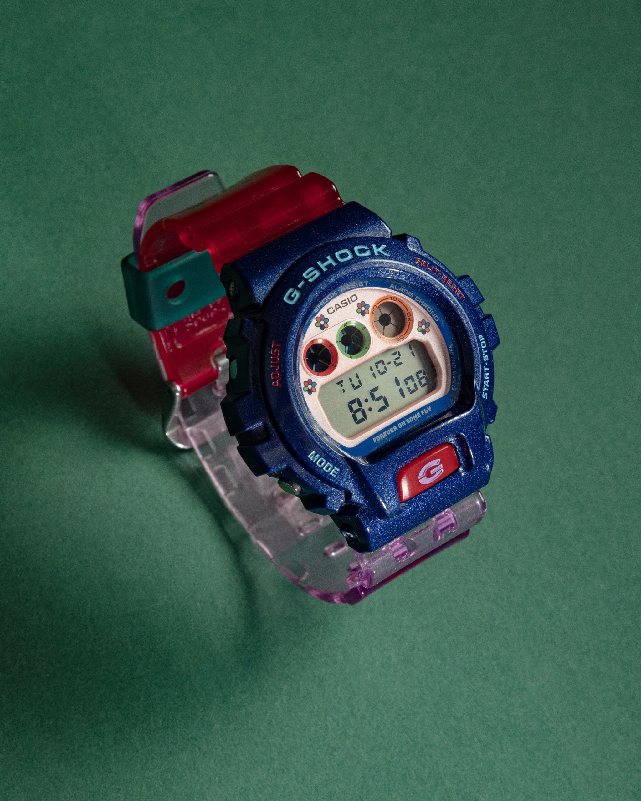 A colorful Casio G-Shock watch with a blue face, red strap, and digital display on a green background.