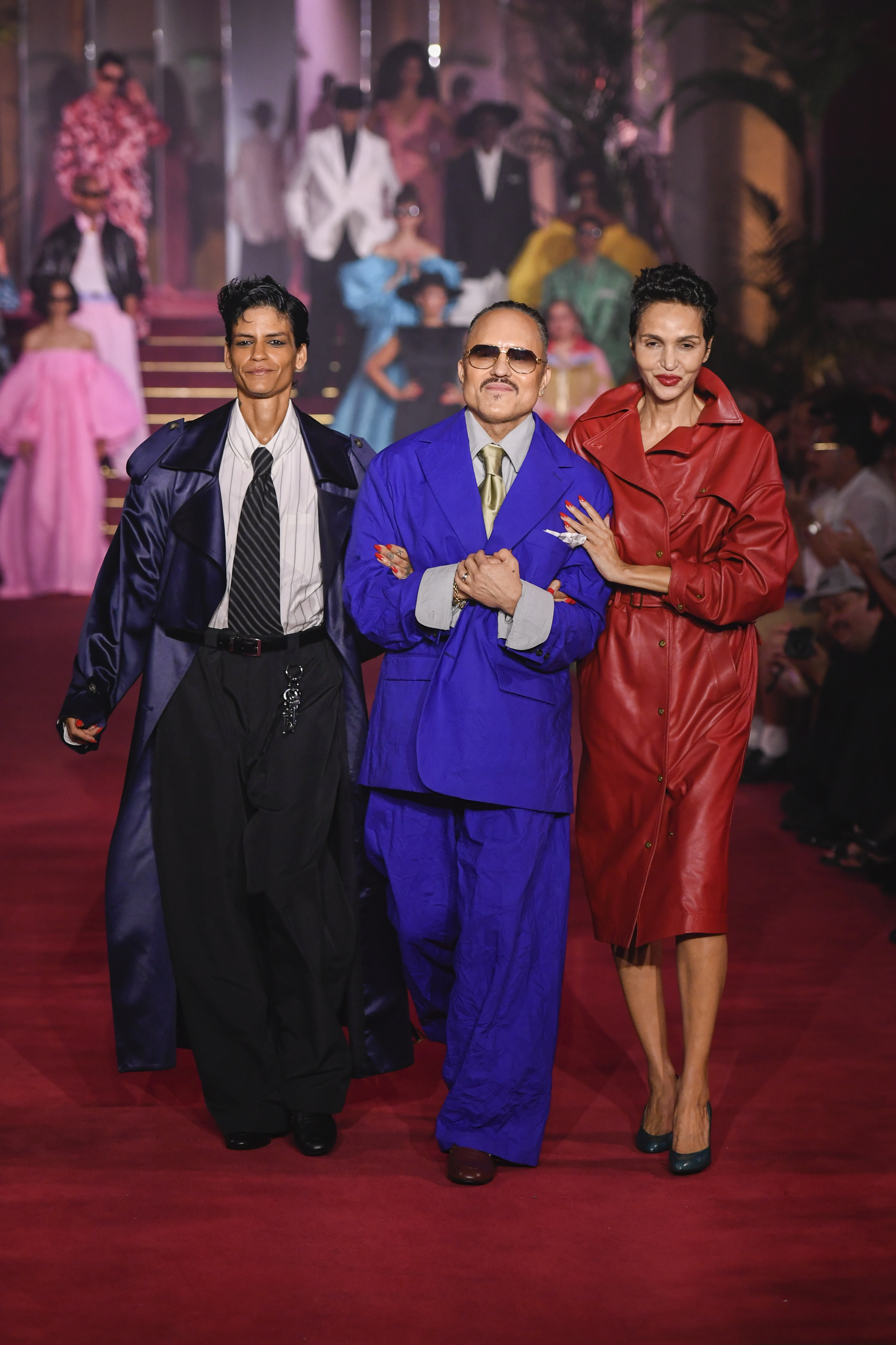 Three people walking on a red carpet runway, dressed in bold, colorful outfits. The background features models and vibrant lighting.