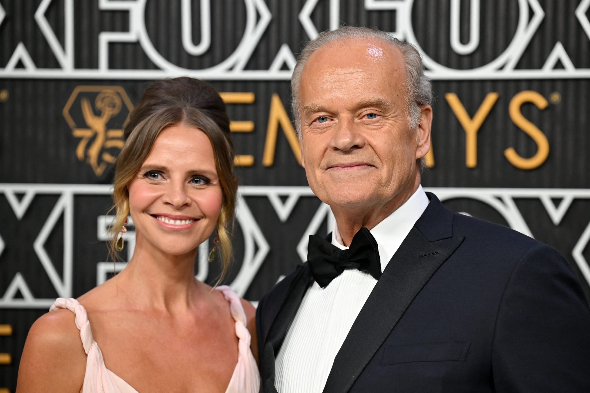 Kelsey Grammer and his wife Kayte Walsh posing at an event, with the man in a tuxedo and the woman in a light pink dress, smiling at the camera.
