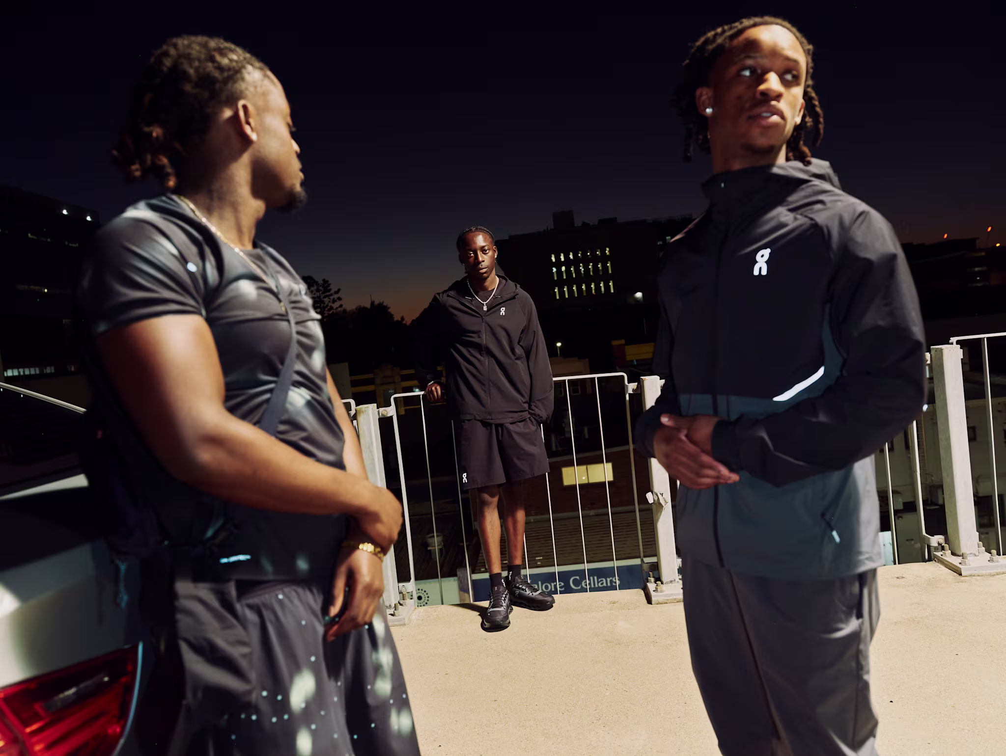 Four individuals in black athletic wear pose against a brick wall under a clear blue sky, with two seated on the wall.