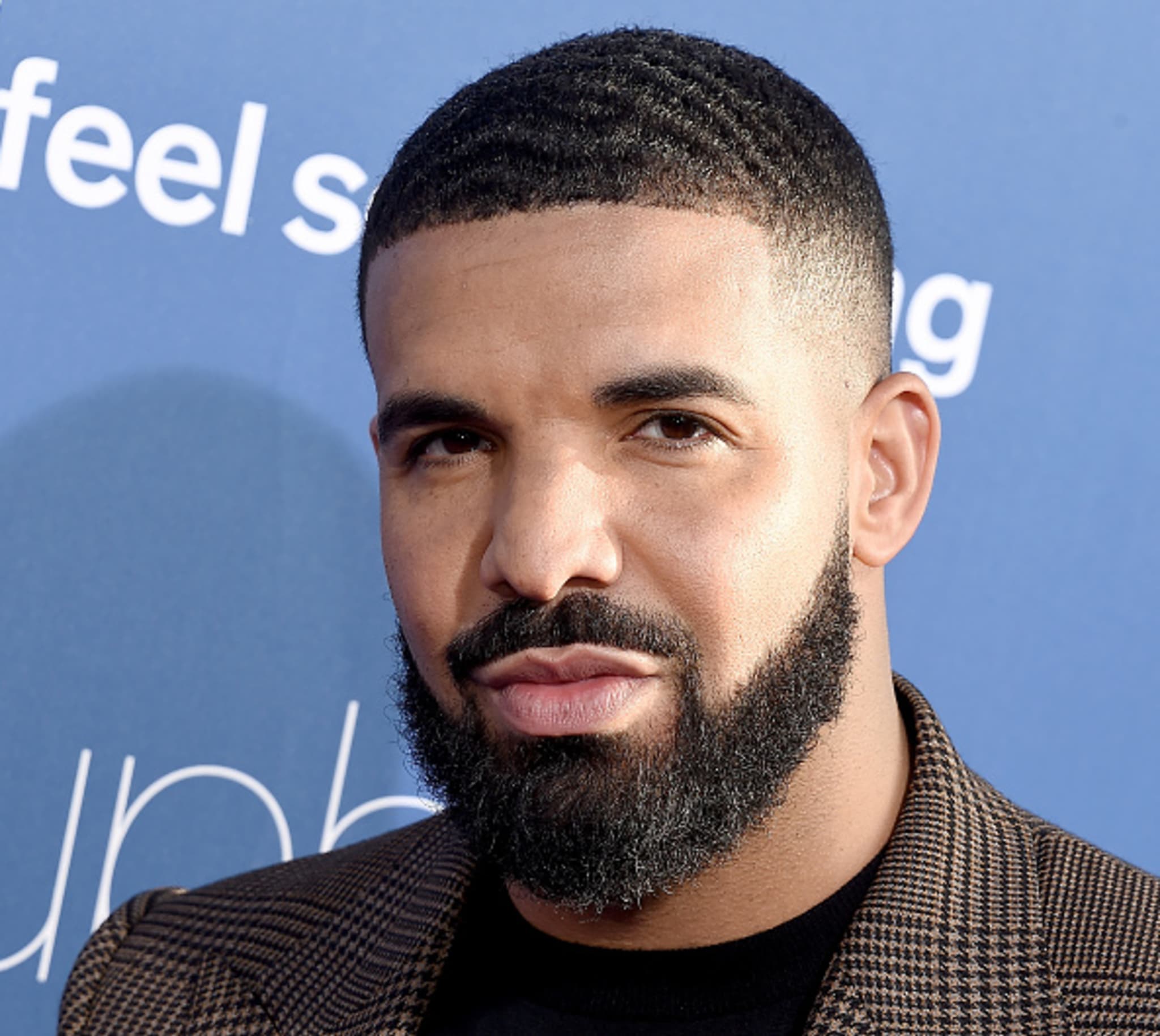 Drake with a neatly trimmed beard and short hair, wearing a brown checkered jacket, standing against a blue background.