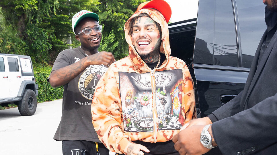 Two people smiling outdoors, one wearing a colorful hoodie and cap, standing near a black vehicle with greenery in the background.