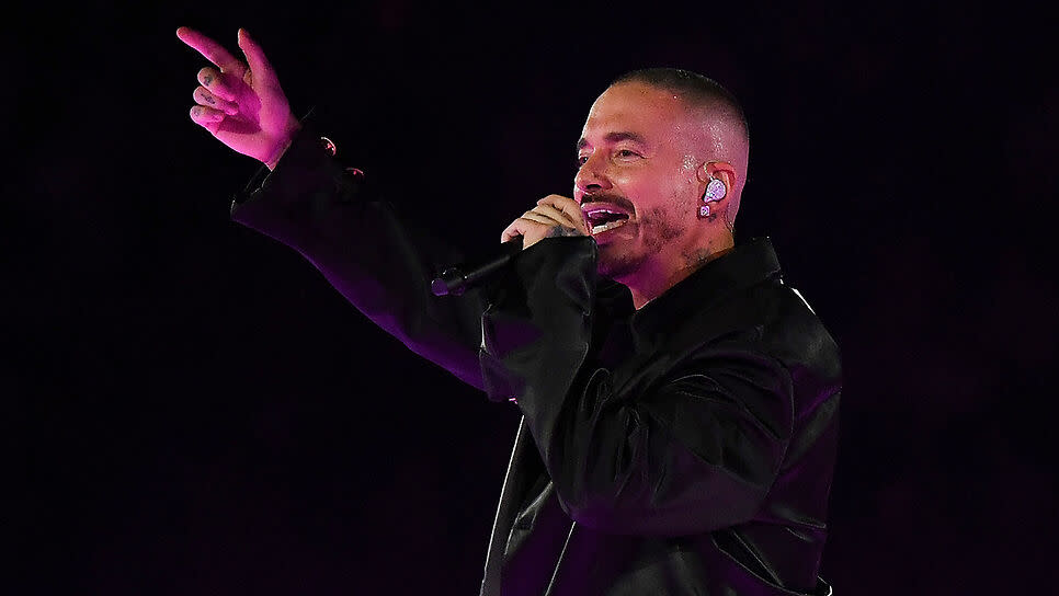 A man in a black jacket is performing on stage, holding a microphone and pointing with his right hand.