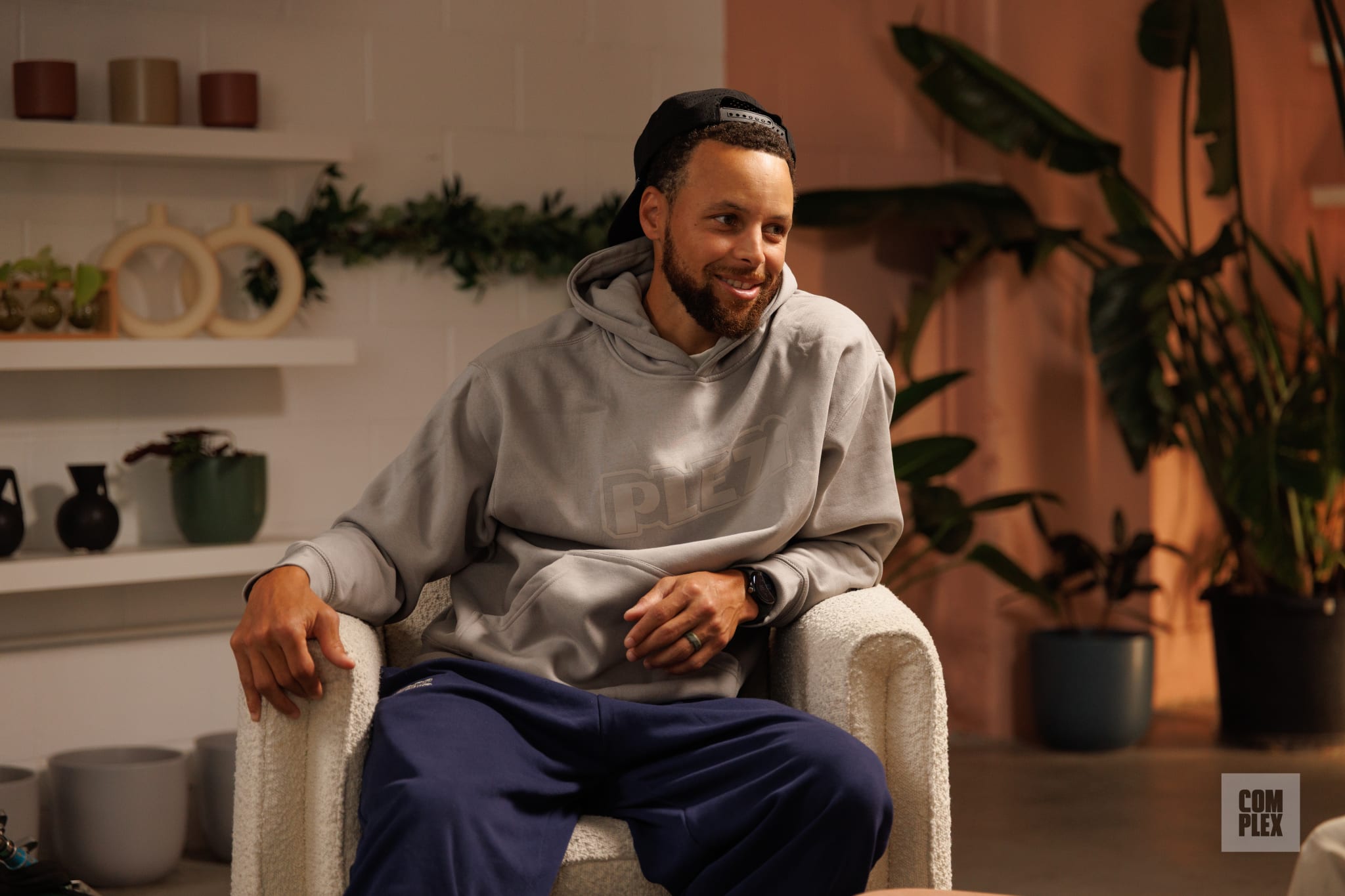Stephen Curry, wearing a gray hoodie and cap, sits in a cozy room with plants and decor.