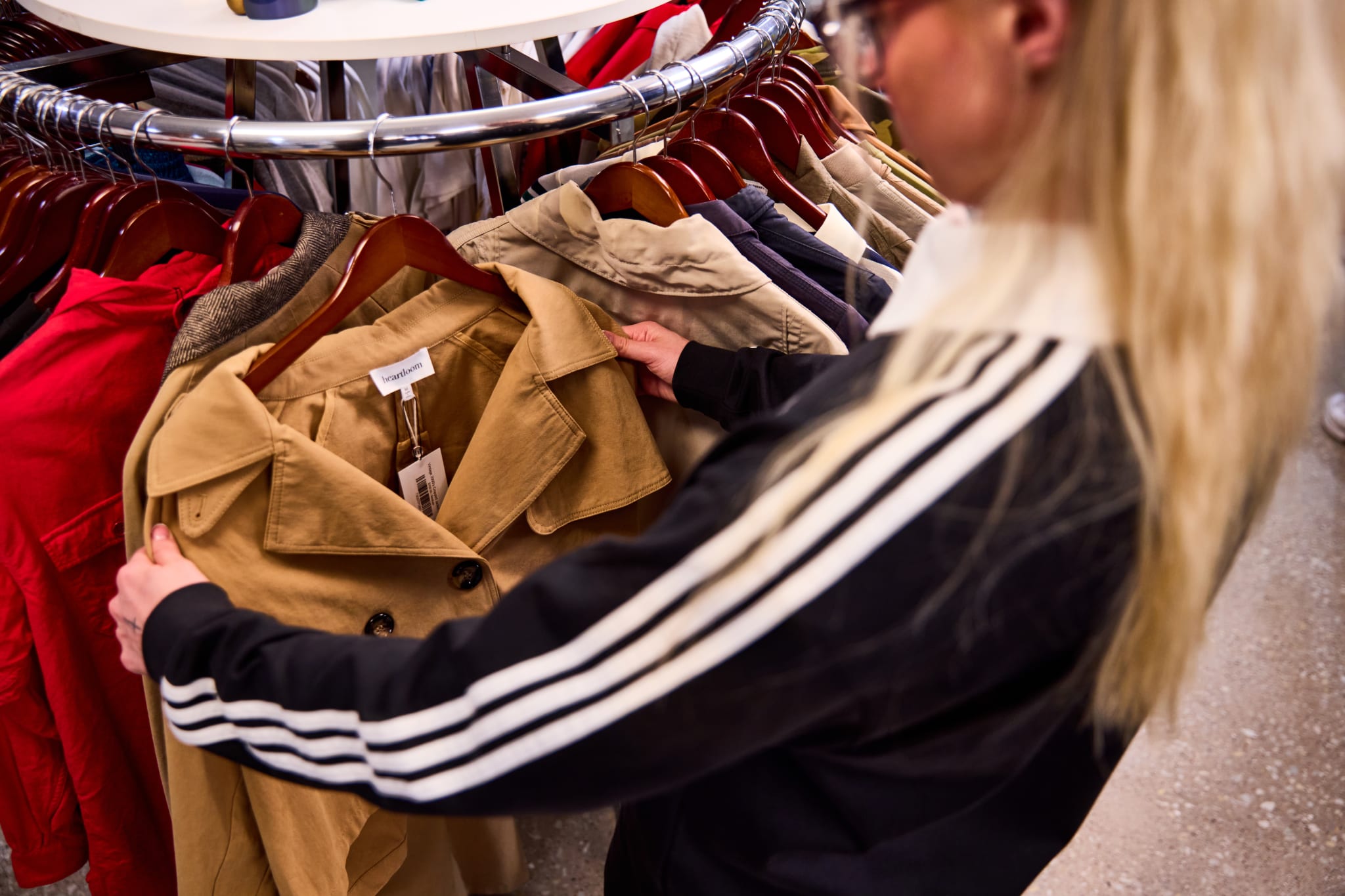 A person with long blonde hair is shopping, holding a tan coat on a rack filled with various colored jackets.