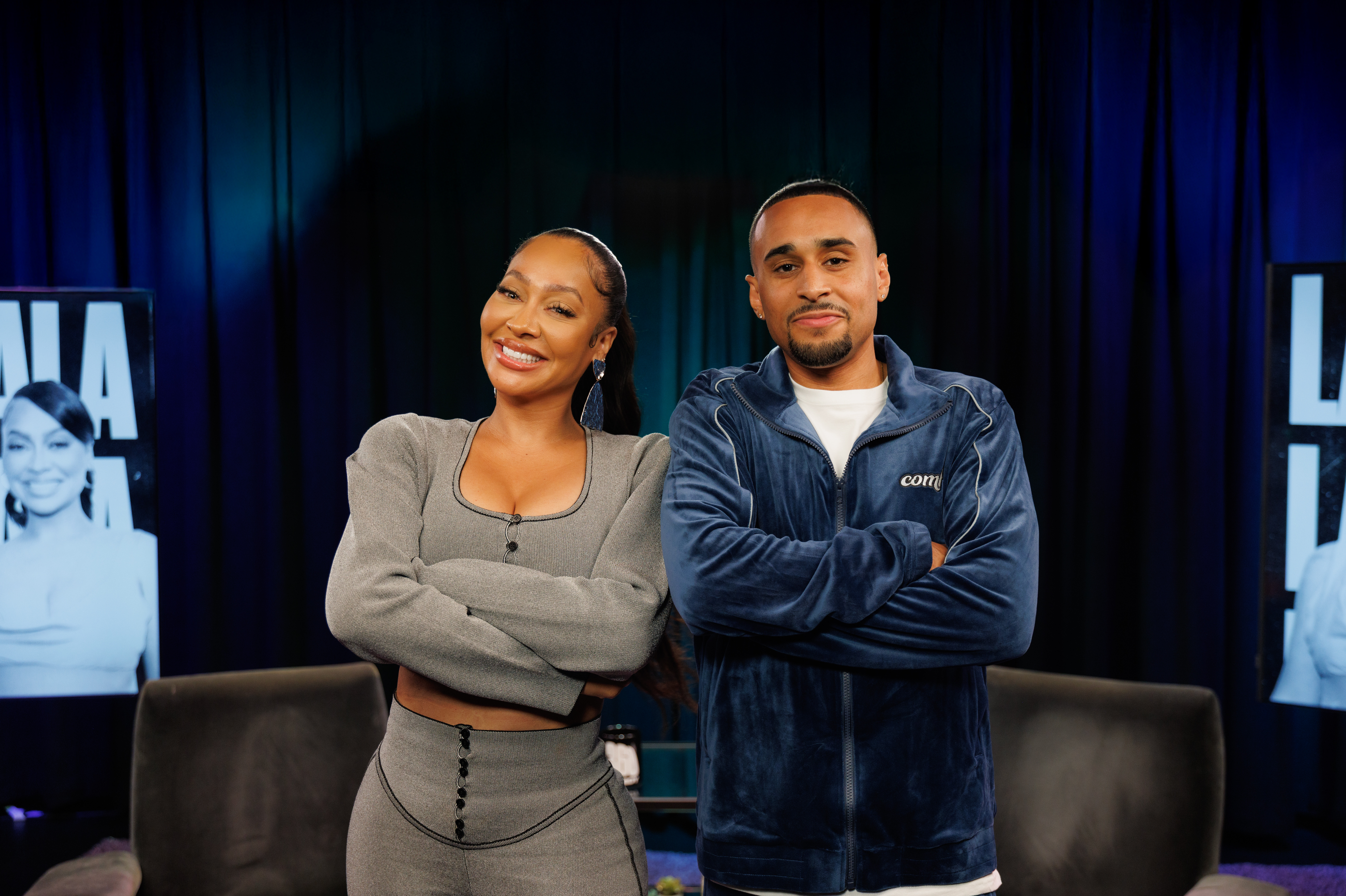 Two people standing with arms crossed, smiling in a studio setting with dark curtains and screens displaying a woman's image.