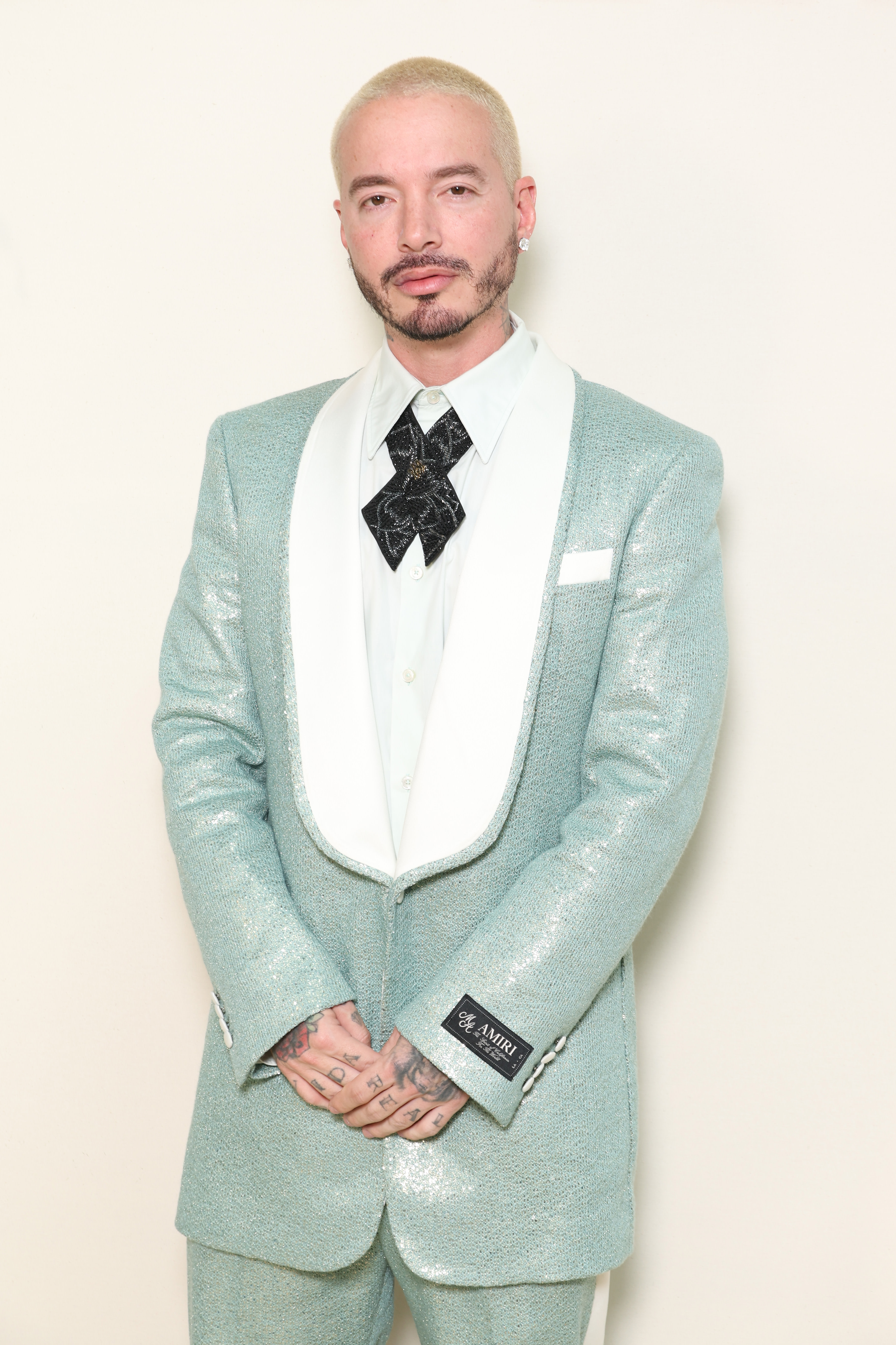 J Balvin in a shimmering light blue suit with a white shirt and black tie, posing against a plain background.