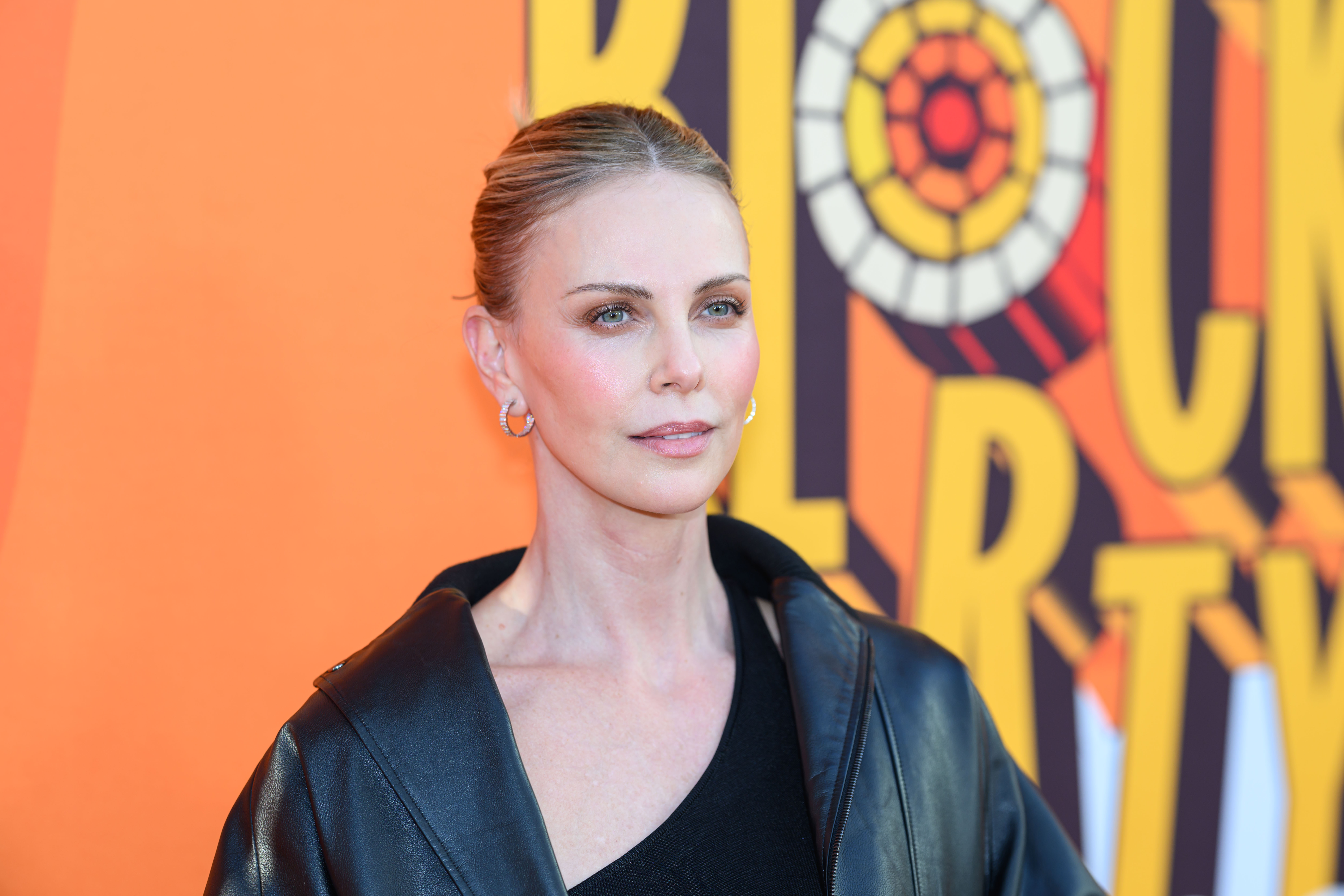 Charlize Theron at an event, wearing a black outfit and hoop earrings, with an orange backdrop featuring bold text and patterns.