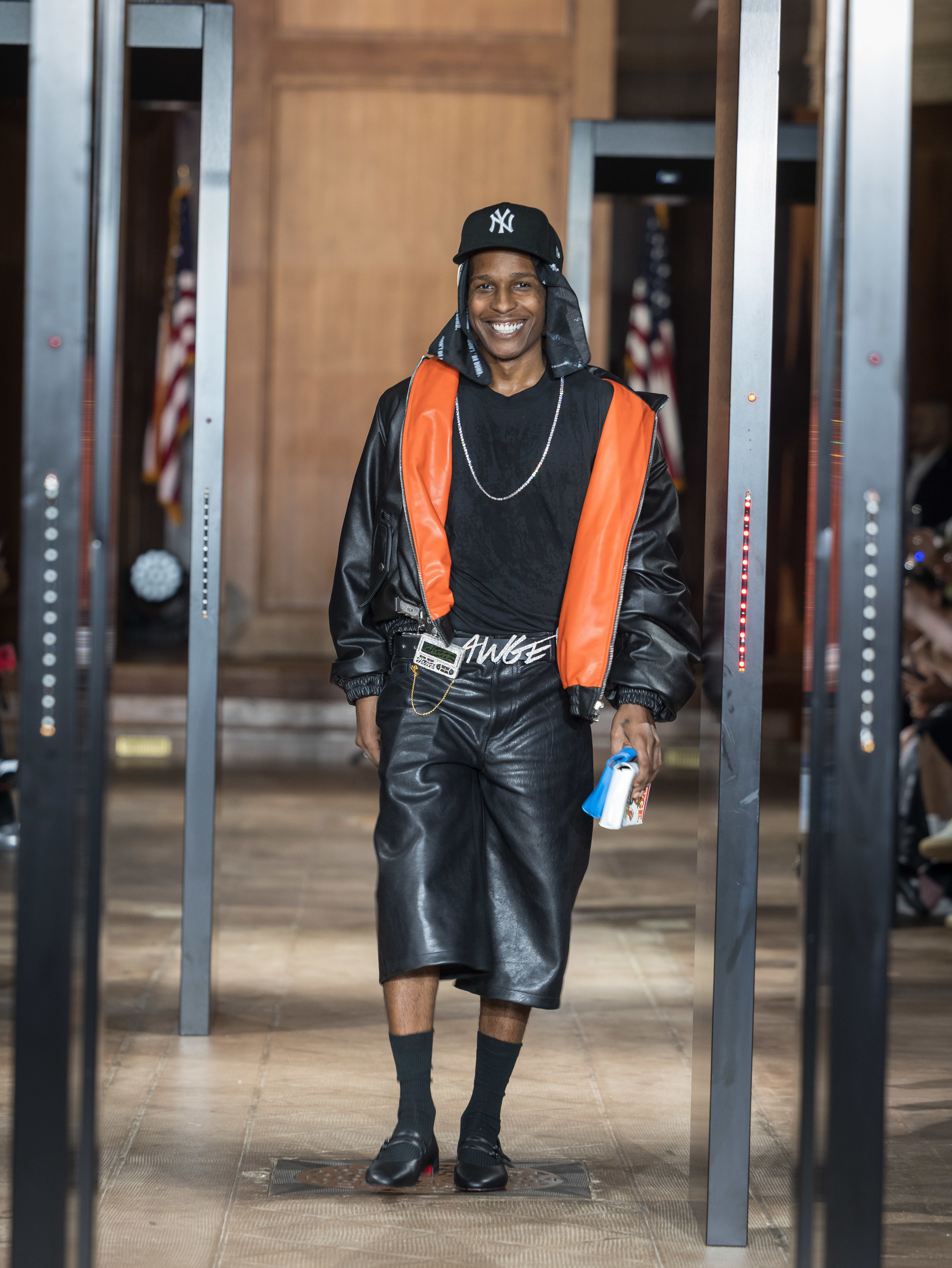 A$AP Rocky walking confidently, wearing a black and orange leather outfit with a New York Yankees cap, smiling.