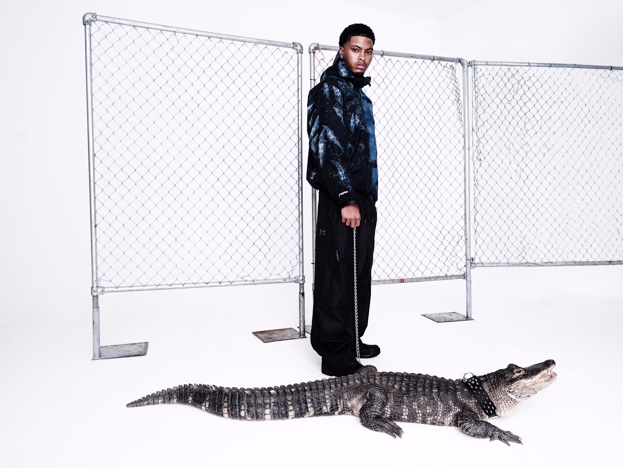 A man in a stylish black and blue outfit stands in front of a chain-link fence, holding a leash attached to an alligator.
