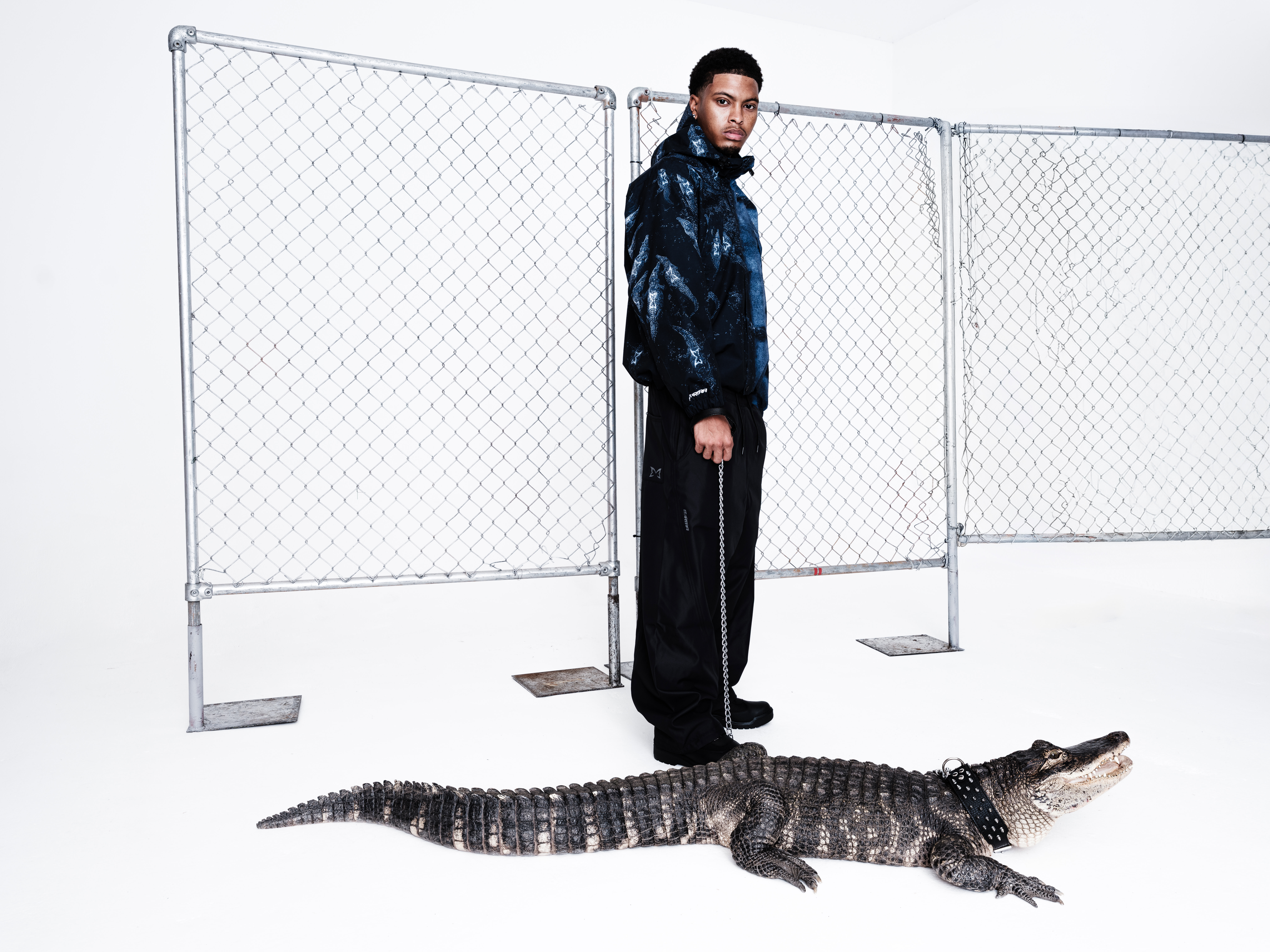 A man in a stylish black and blue outfit stands in front of a chain-link fence, holding a leash attached to an alligator.