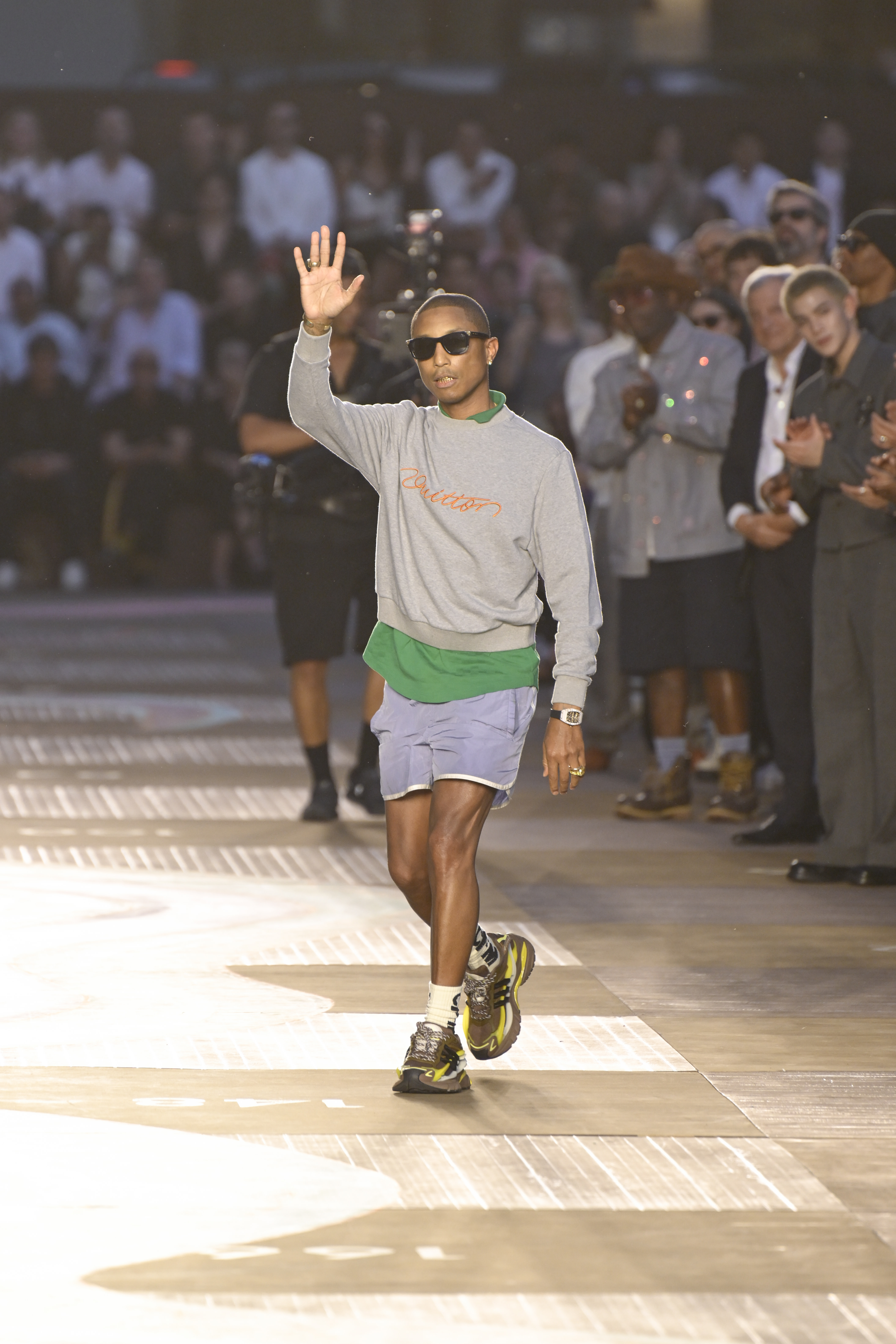 Pharrell Williams waves on a runway, wearing sunglasses, a gray sweater, and shorts, with a crowd applauding in the background.