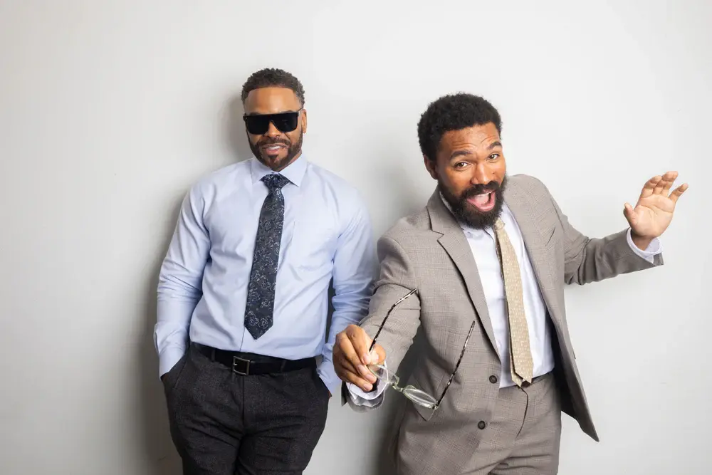 Two men in suits, one wearing sunglasses, posing playfully against a plain background.